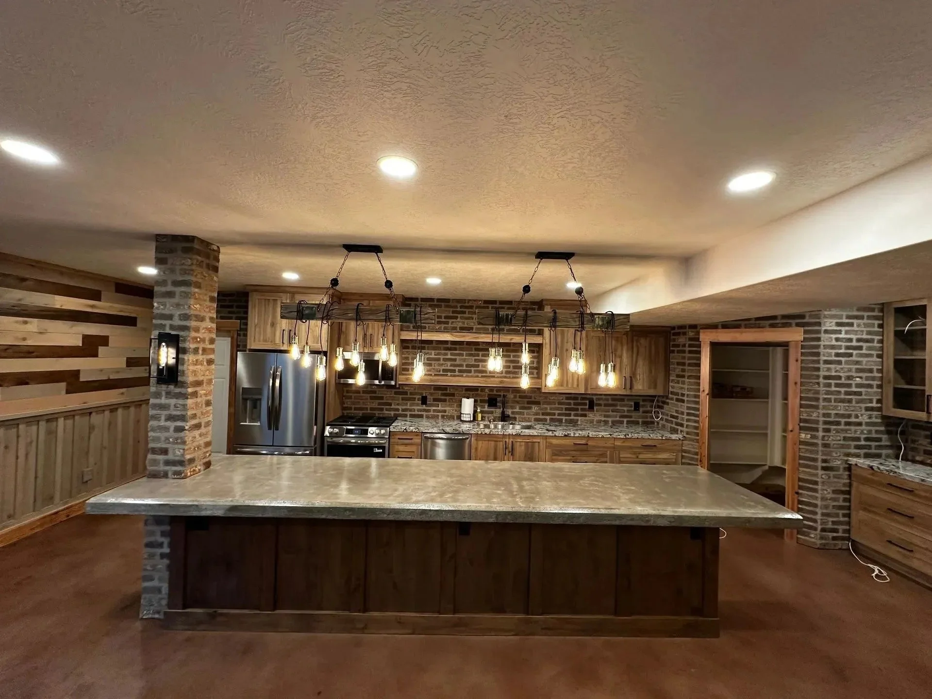 Modern kitchen with a large center island, brick accents, wooden cabinets, hanging pendant lights with Edison bulbs, stainless steel refrigerator, and wood-paneled walls.