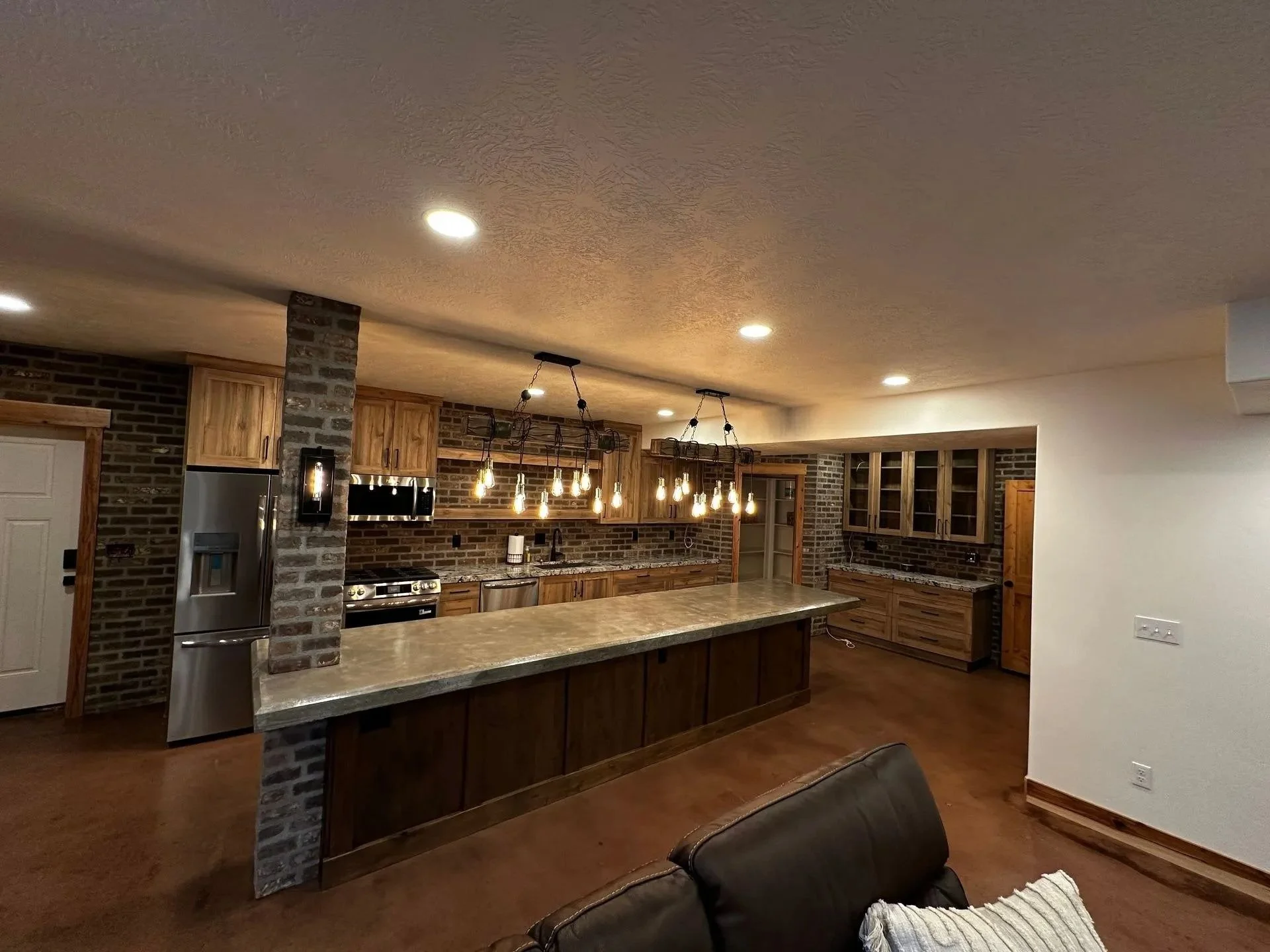 Interior view of a modern kitchen with wooden cabinets, stainless steel appliances, and a large stone counter. The ceiling features recessed lighting and hanging Edison bulbs, with a brick wall and additional cabinetry in the background.