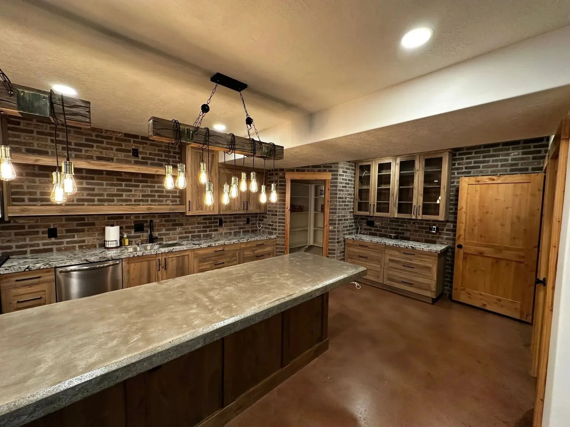 A spacious kitchen with a large island, wooden cabinets, brick walls, and a hanging light fixture with multiple Edison bulbs.