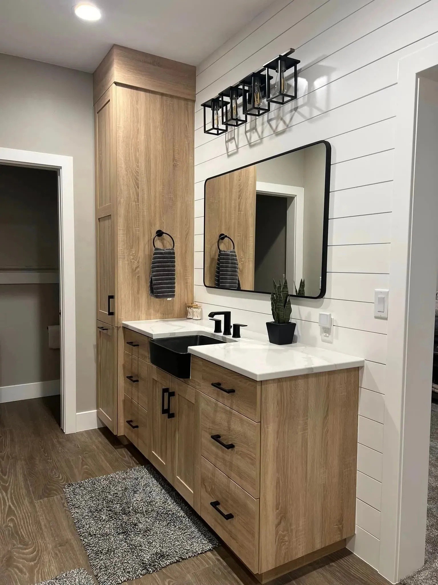 Modern bathroom with natural wood cabinets, black hardware, a large mirror, black sink, black faucet, a small plant, and a black geometric ceiling light fixture. The walls are white with horizontal siding, and there is a gray textured rug on the floo