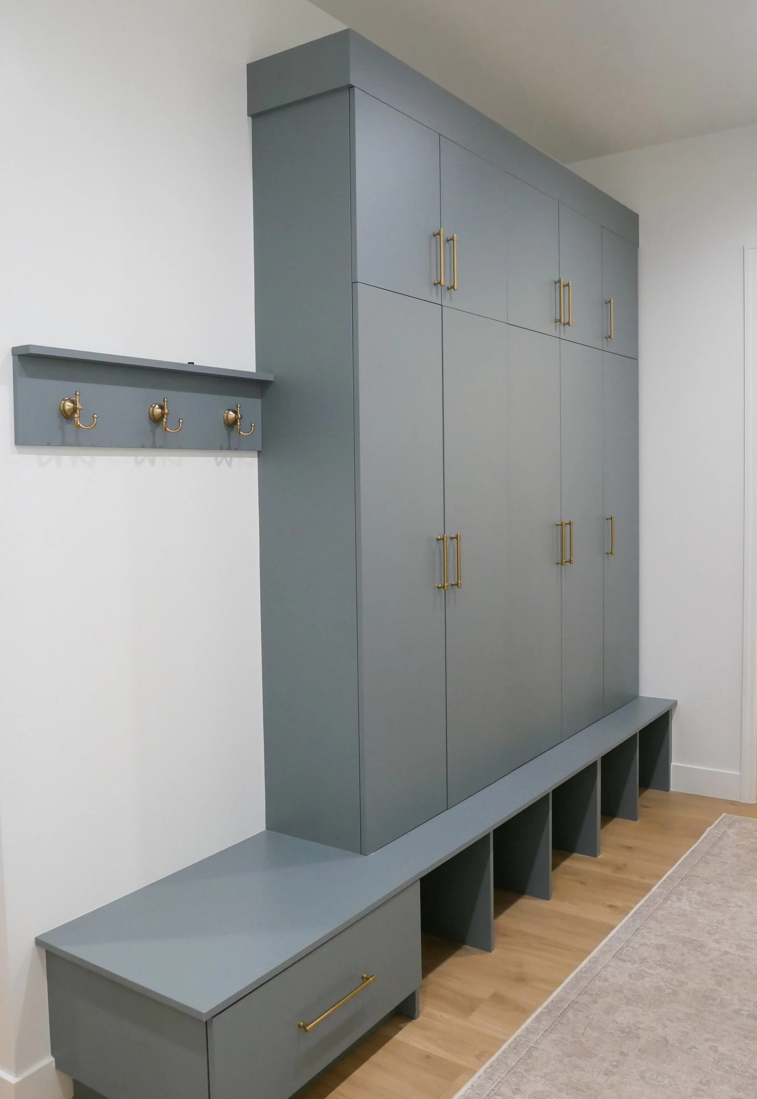 A built-in storage cabinet in a modern home, painted in muted blue, with gold handles, positioned against a white wall with a small shelf and hooks nearby.