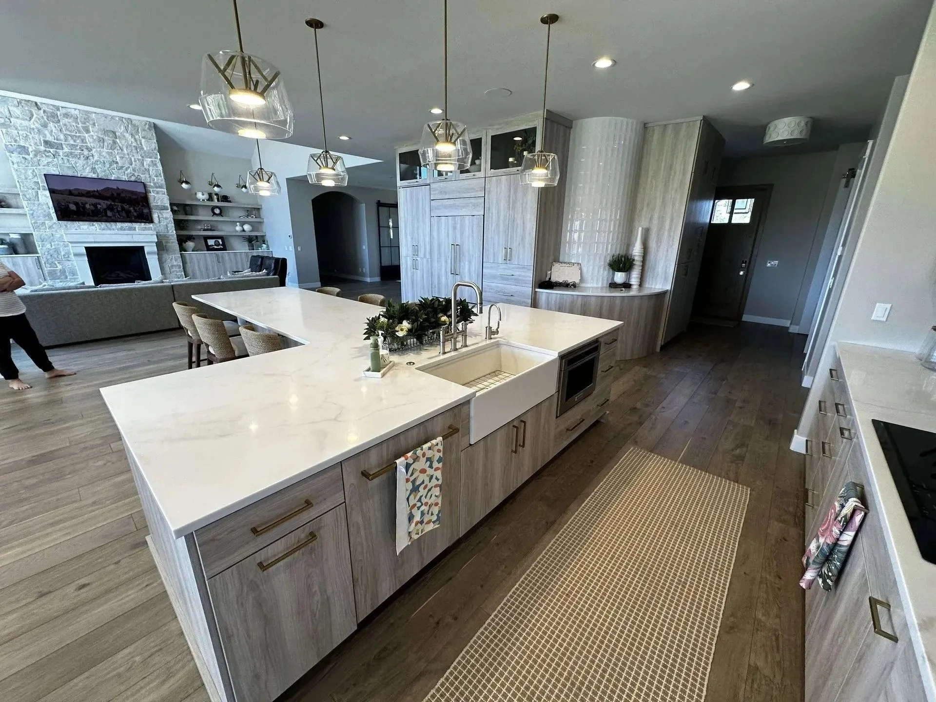 Modern kitchen with a white marble island, light wood cabinets, and pendant lighting, adjacent to a living room with a stone fireplace and TV.