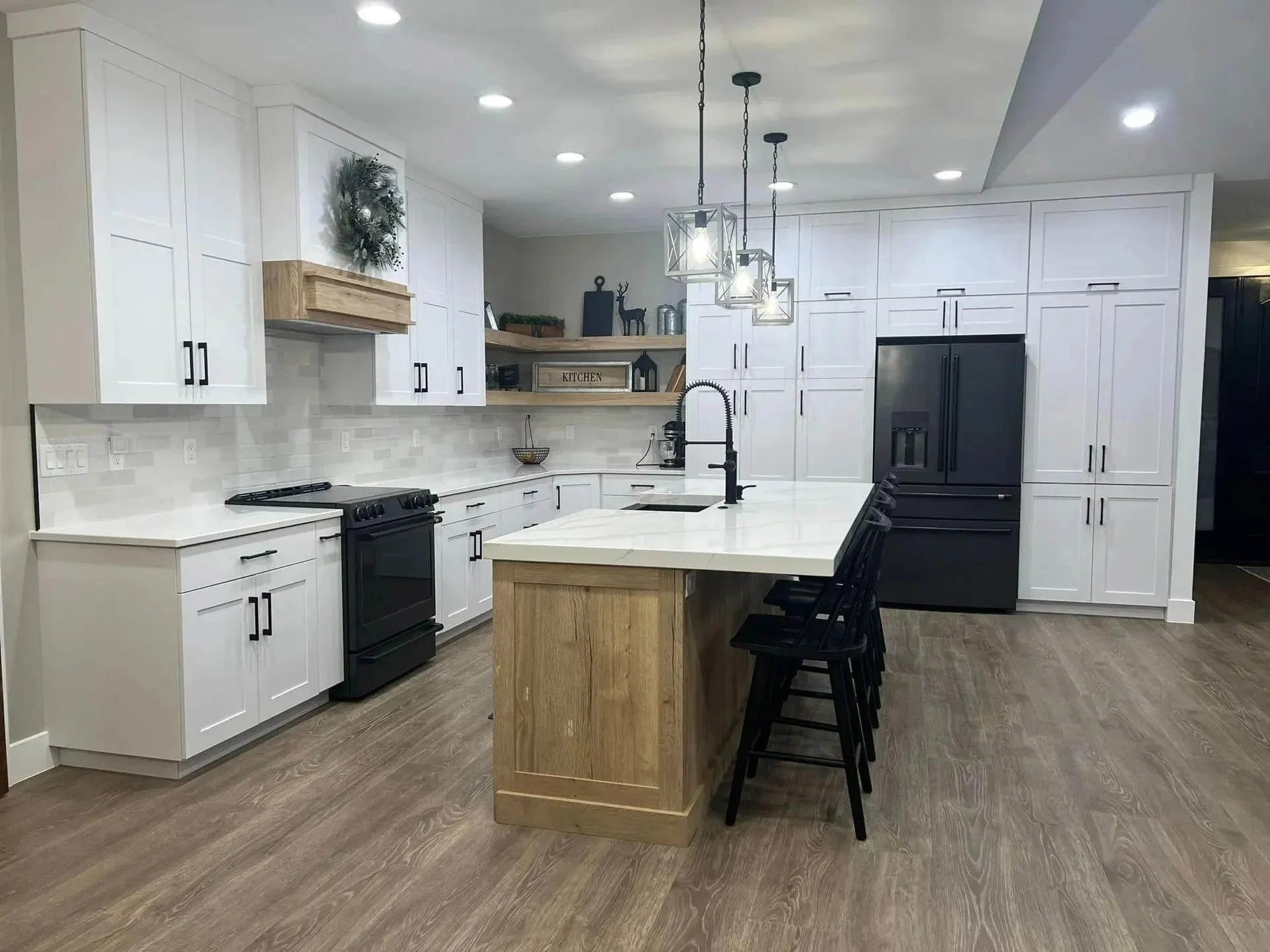 Modern kitchen with white cabinets, black appliances, island with a wooden base, and black bar stools, decorated with holiday greenery and black decor.