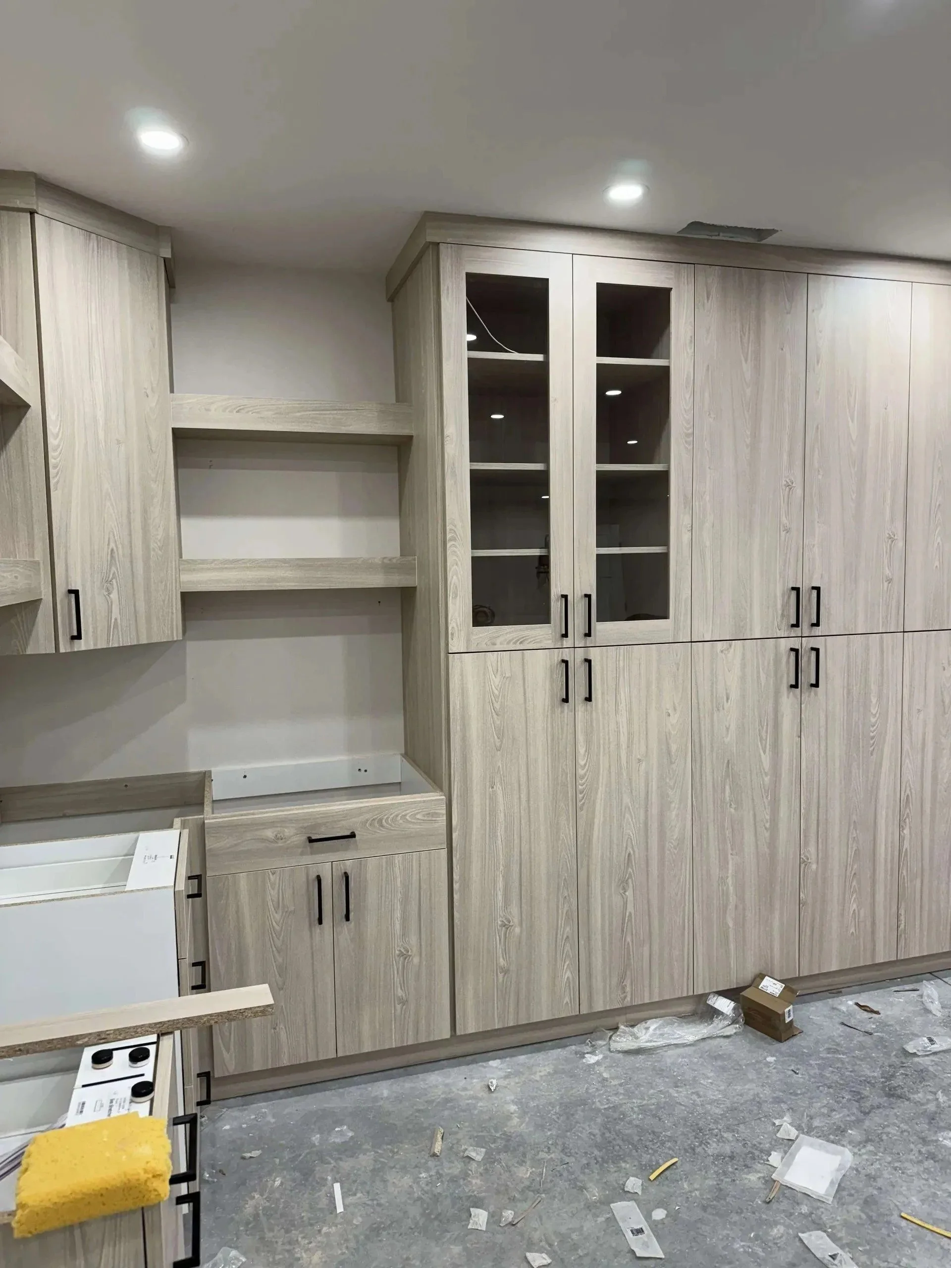The image shows a room under construction or renovation, with light-colored wooden cabinets installed on the walls. There is a messy floor with debris, plastic wrap, a yellow cleaning cloth, and a small cardboard box. The ceiling has recessed lightin