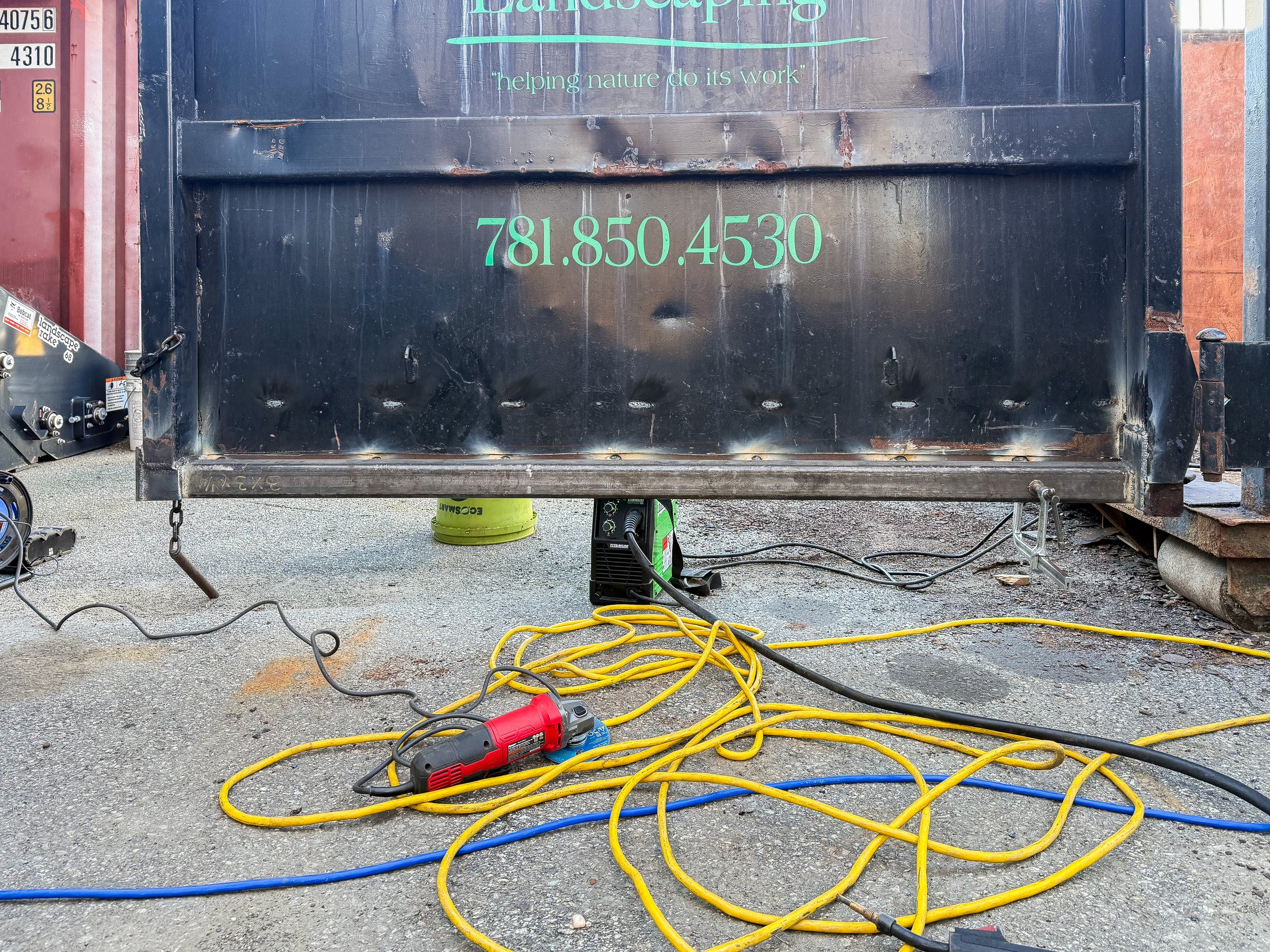 A construction or landscaping setup featuring a large black metal container with the word 'Landscaping' and a phone number written in green. There are yellow and blue extension cords and power tools on the ground in front of the container, with some electrical equipment and a green bucket visible underneath.