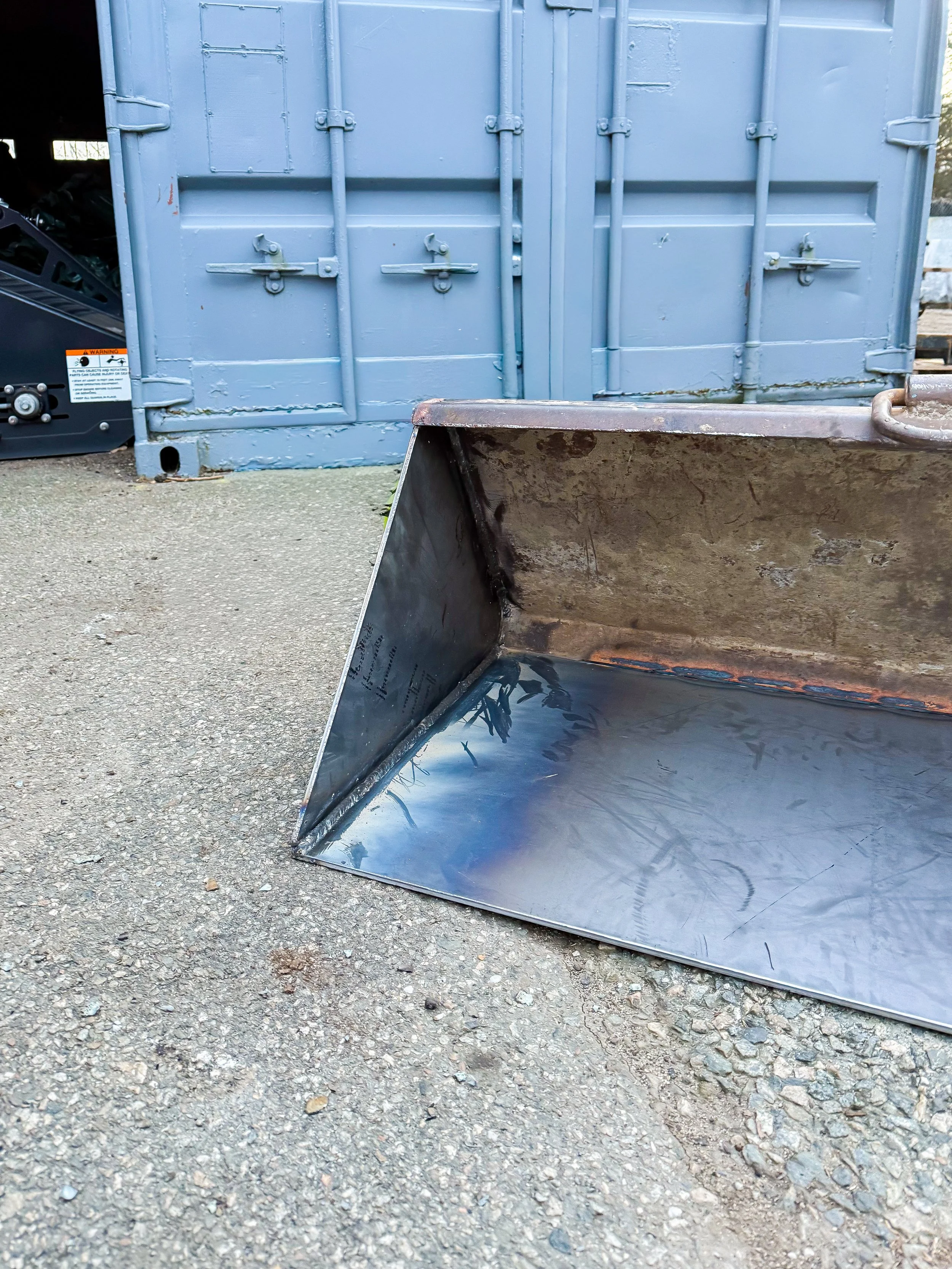 A metal scoop or shovel in front of a closed blue shipping container or storage unit, with gravel or rough concrete ground underneath.