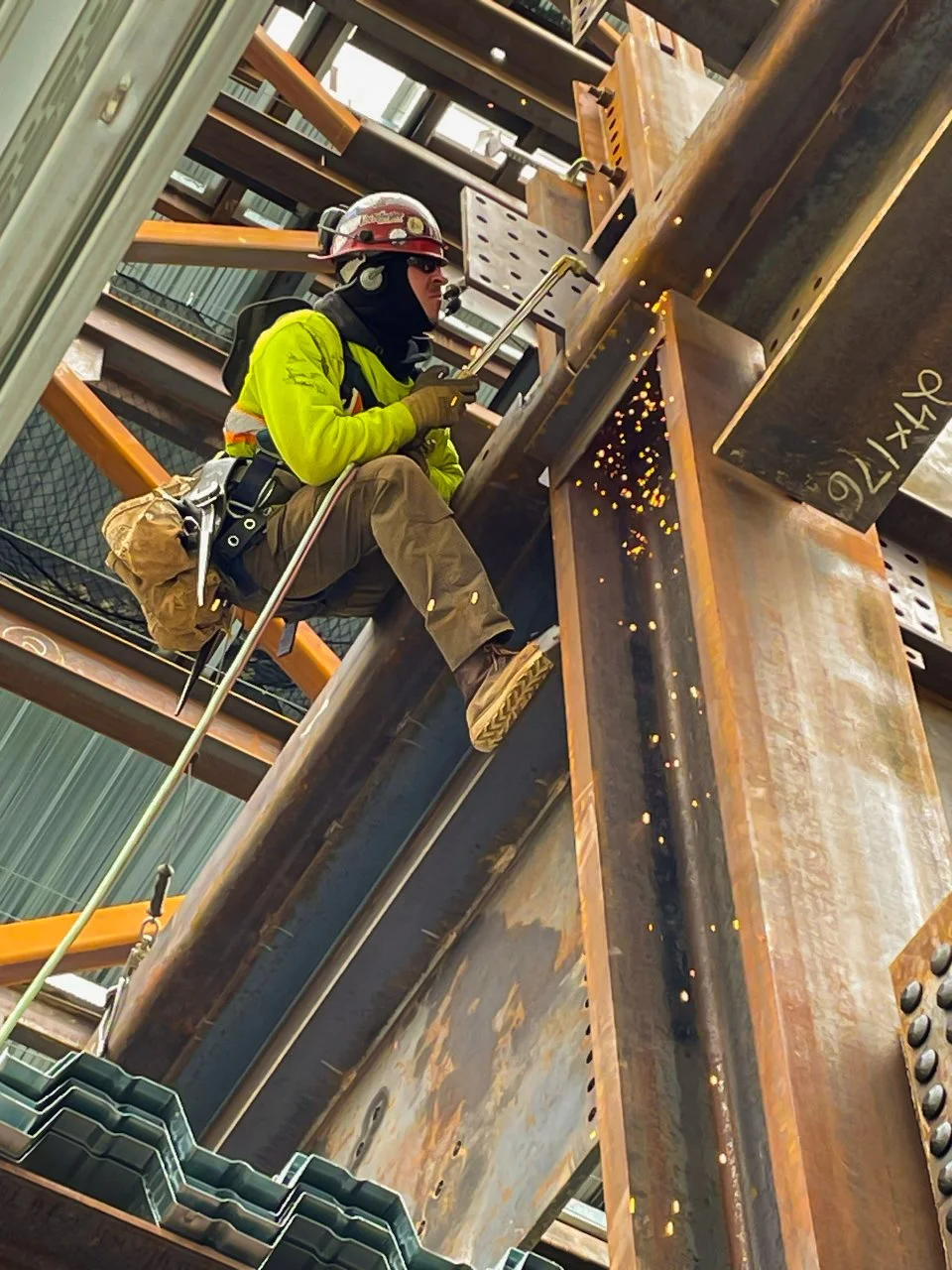 Construction worker wearing a yellow safety jacket, helmet, and headphones, welding metal beams in an industrial construction site, with sparks flying from the welding process.