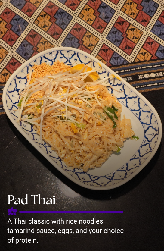 A plate of Pad Thai with rice noodles, bean sprouts, a lemon wedge, and crushed peanuts on a blue and white patterned dish, placed on a table surface.