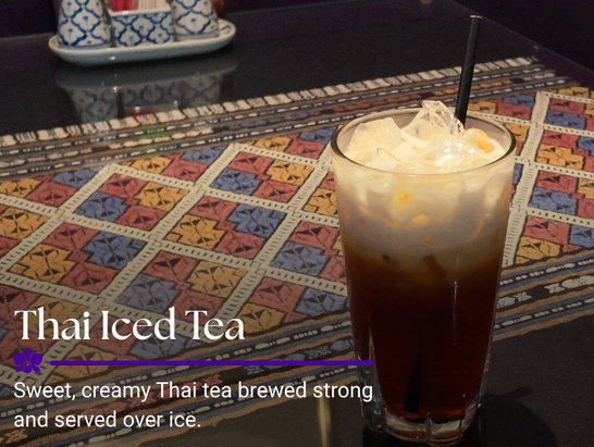 A tall glass of Thai iced tea with ice and cream on top, placed on a decorative tablecloth at a restaurant.