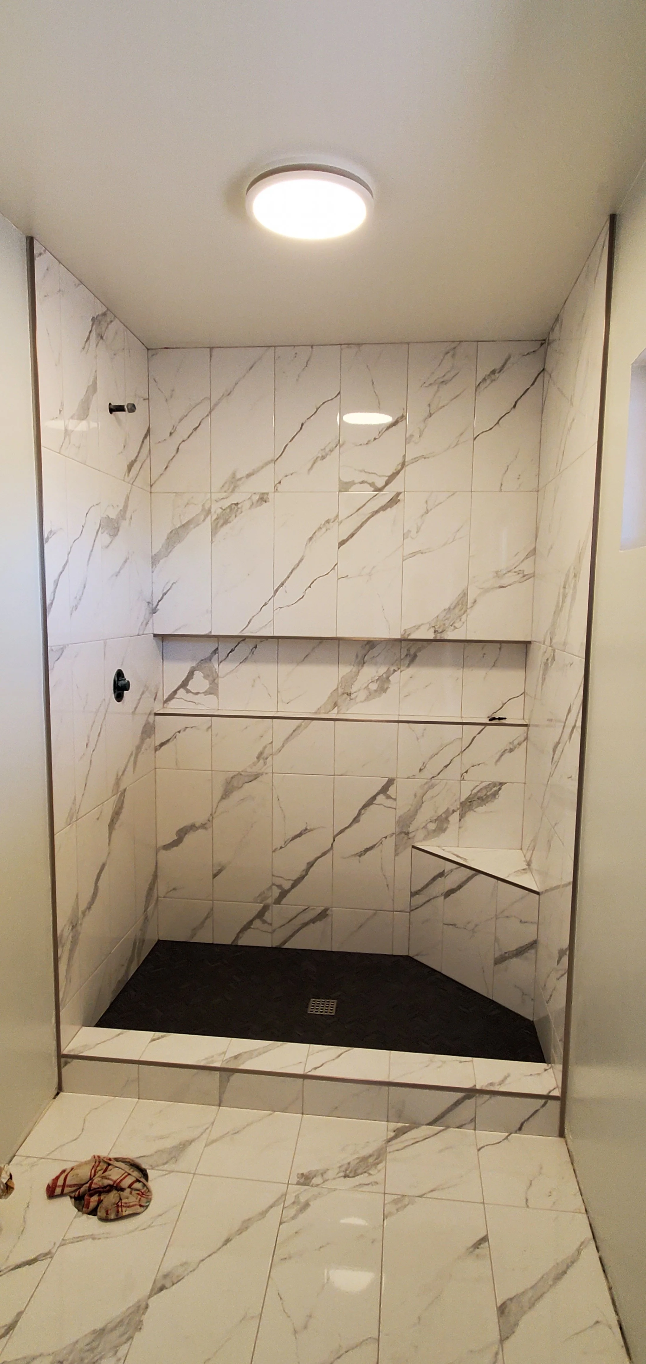 A walk-in shower with white marble tiles featuring gray veining, a built-in bench, and a drain on the dark floor. The shower has no visible fixtures and a ceiling-mounted light.