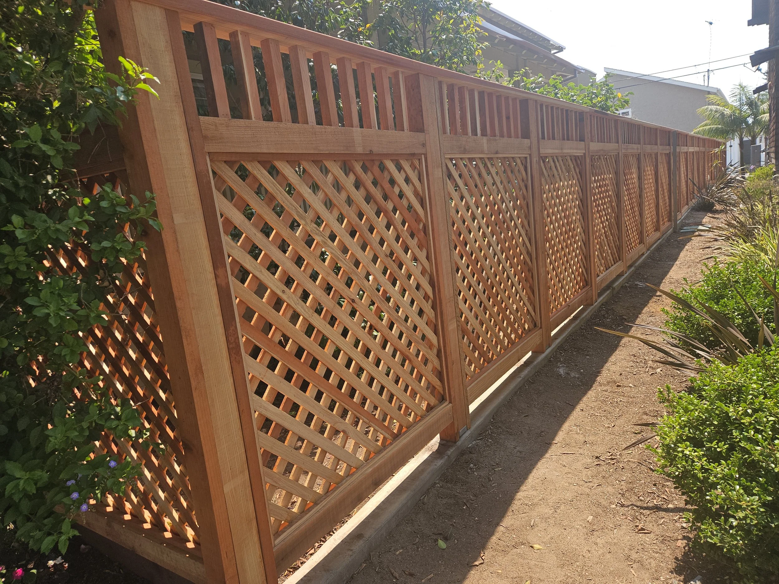 Long wooden lattice fence along a garden walkway, with plants and bushes on the side, under bright sunlight.