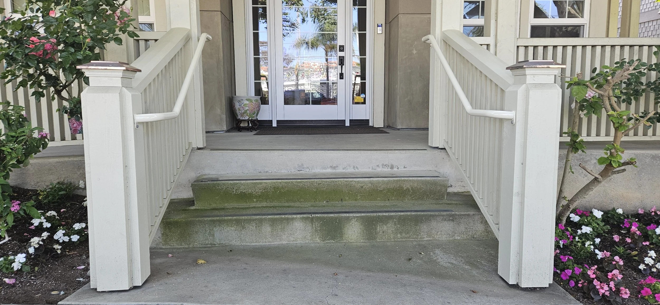 Front porch of a house with stairs, white railings, and a glass door, surrounded by flowering plants and trees.