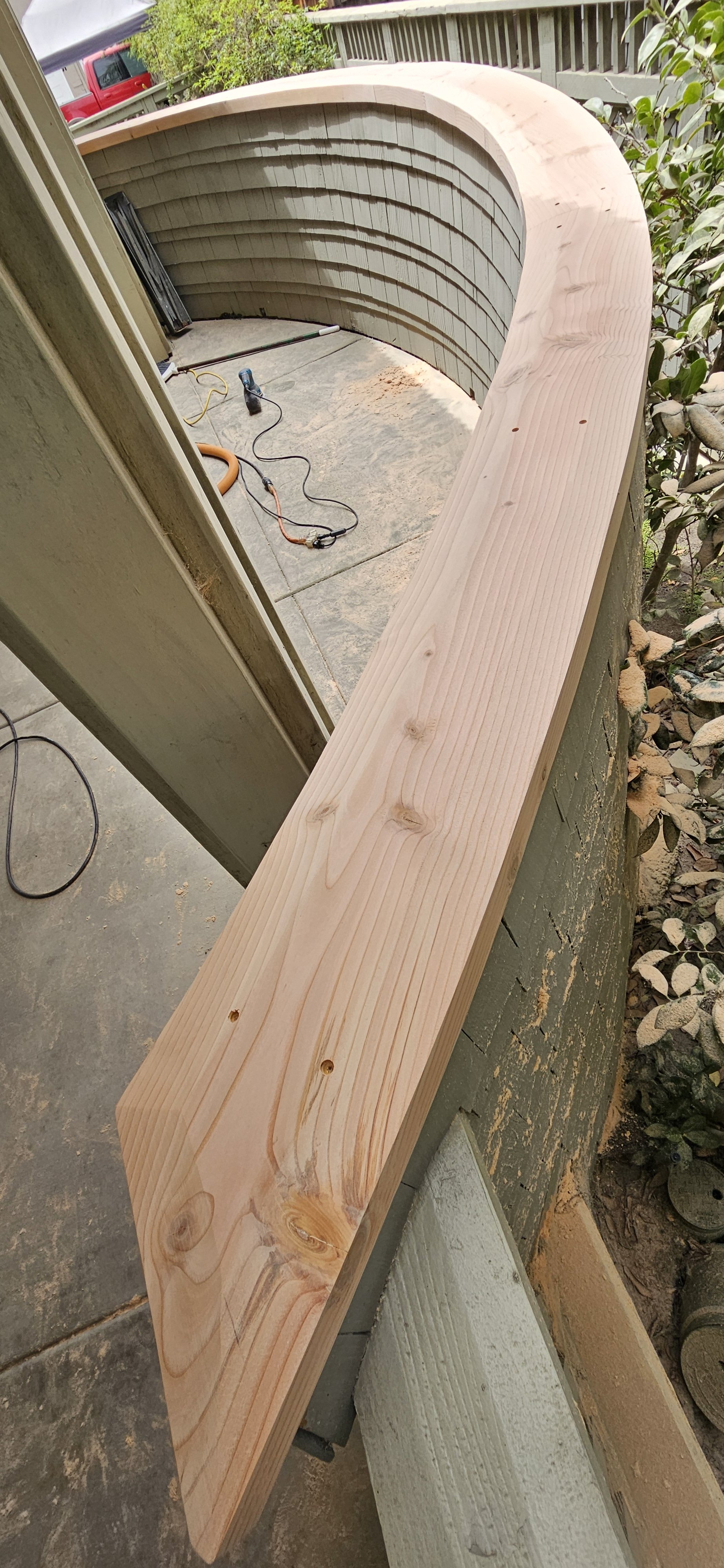 A piece of shaped, unfinished wood rests on the edge of a wall outside, with construction tools and materials nearby.