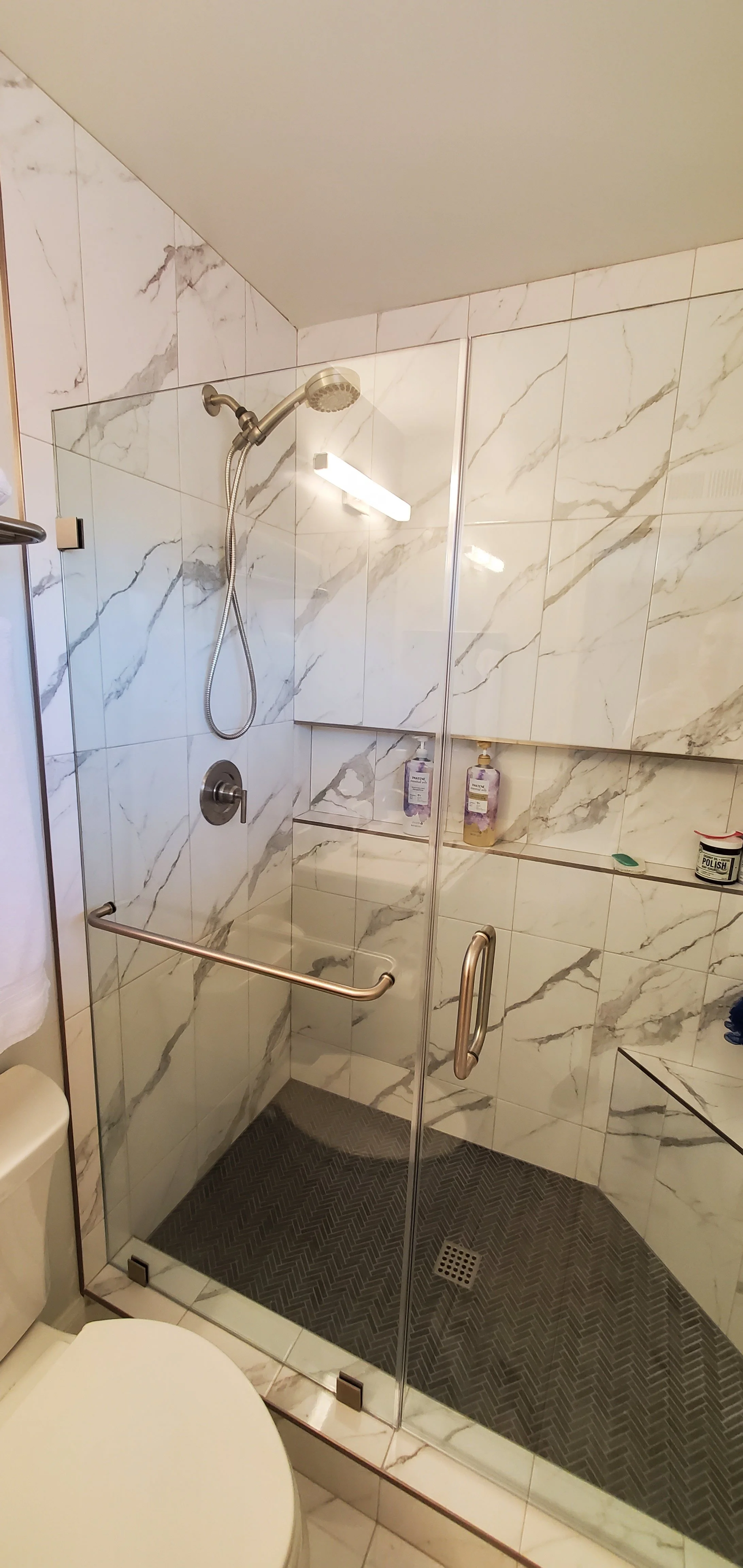 A walk-in shower with marble tile walls, a handheld shower head, a glass door with metal handles, built-in shelves with soap and shampoo bottles, and a dark textured floor.