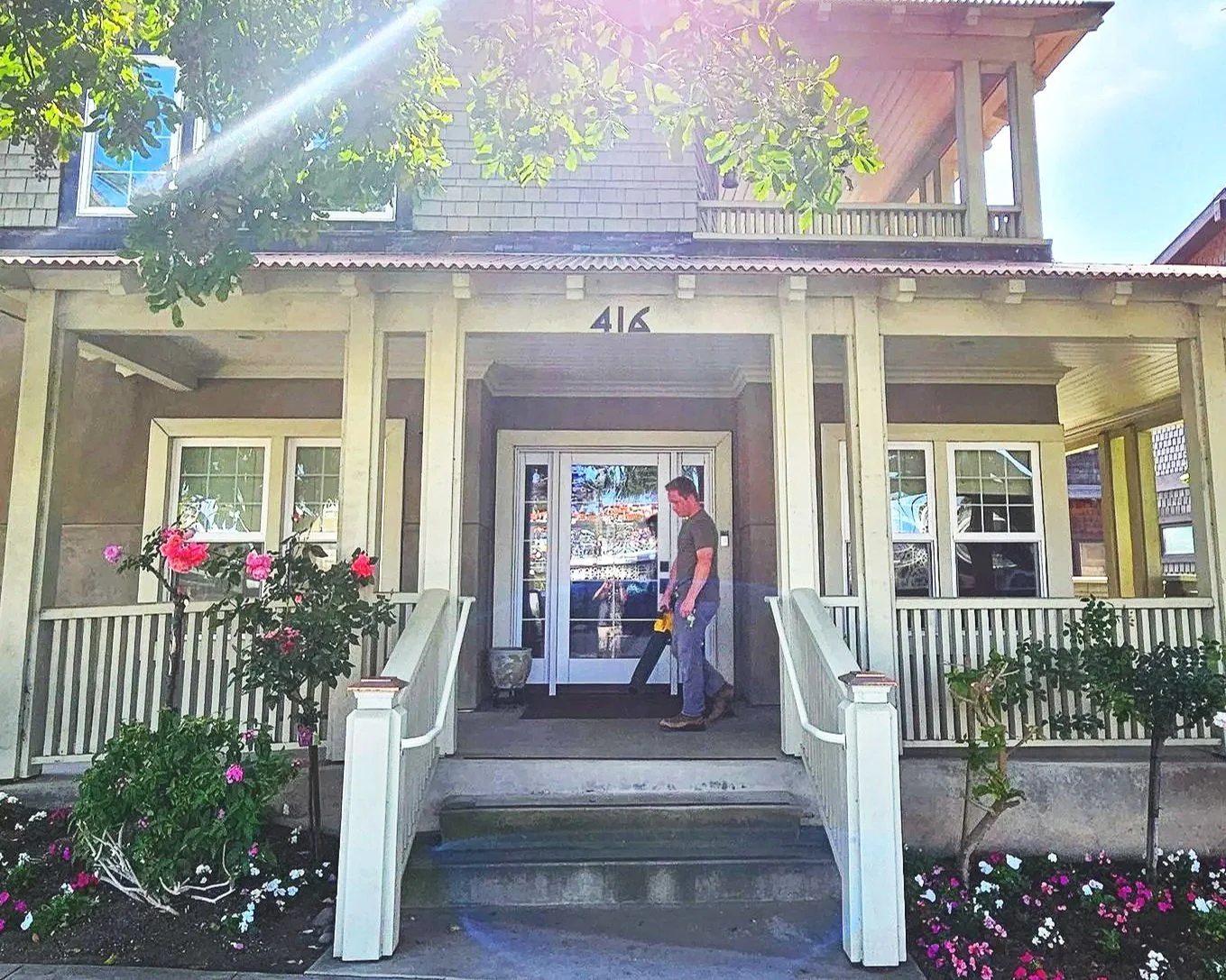 A person standing on the steps of a house with the number 416, holding a saw, with flowers and greenery in front and a sunny sky above.