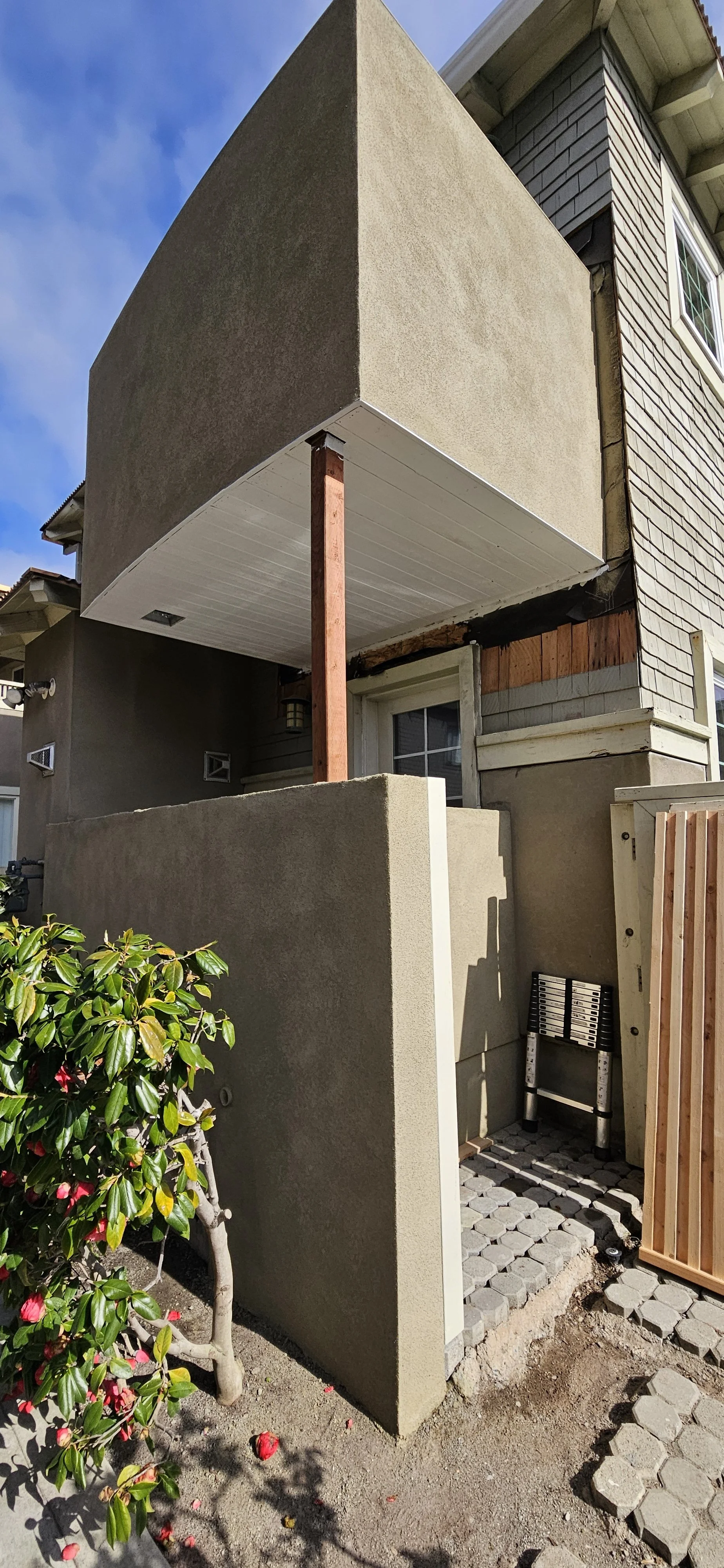 Outside of a building under construction, showing a small balcony with beige stucco walls and a wooden support post, with a bush and a pathway made of hexagonal pavers.
