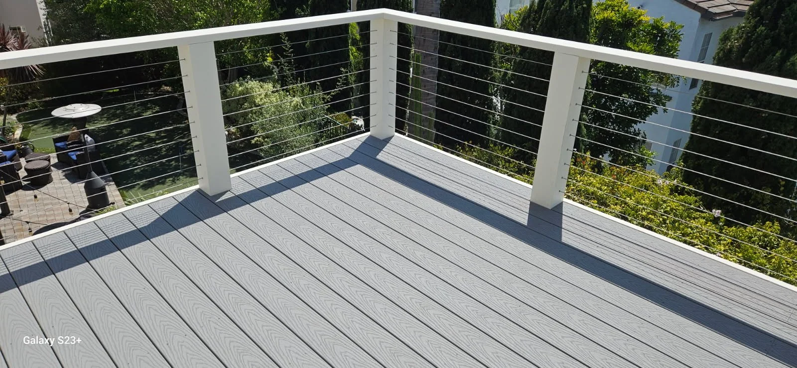 View of a balcony with gray wooden flooring and white railing, cables, overlooking a garden with trees, patio furniture, and a person sitting under a white umbrella.