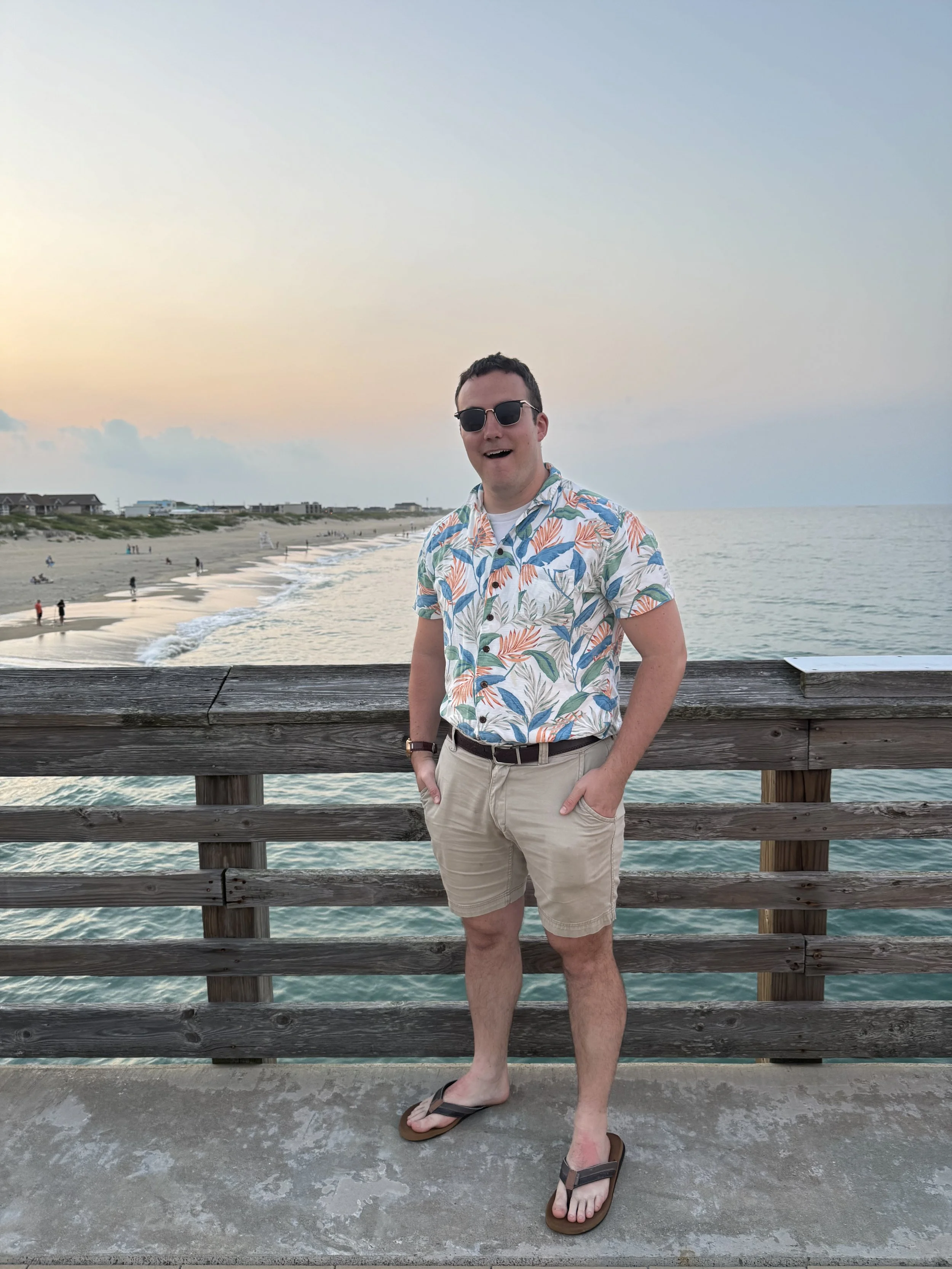 A man wearing a floral Hawaiian shirt, beige shorts, sunglasses, and flip-flops standing on a pier with an ocean behind him. The sky is partly cloudy at sunset, with a beach and shoreline visible in the distance.