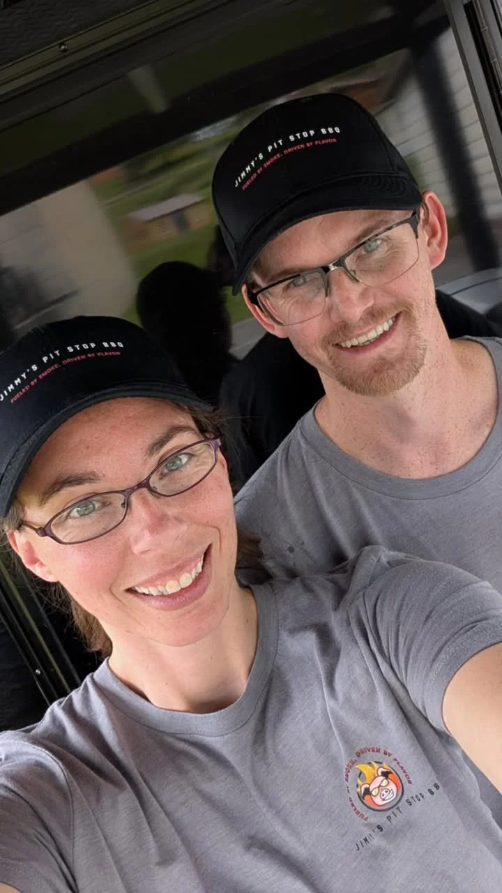 Two smiling people wearing gray T-shirts and black baseball caps with the text "Jimmy's Pit Stop BBQ" on them, taking a selfie outside.