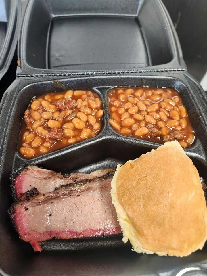 A black compartmentalized food container with baked beans, a piece of cooked steak, and a bread roll.