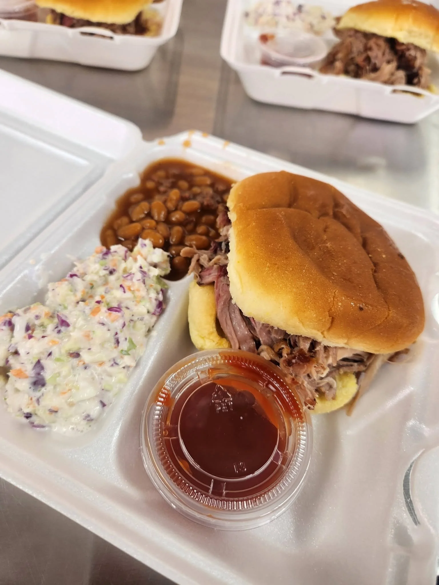 Barbecue sandwich with shredded meat on a bun, served with baked beans, coleslaw, and a side of barbecue sauce.