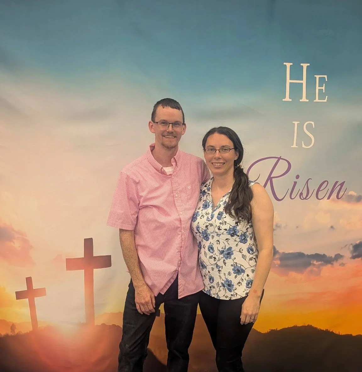 A smiling couple standing in front of a backdrop with crosses and a sunset, with the words "He is Risen" written on it.