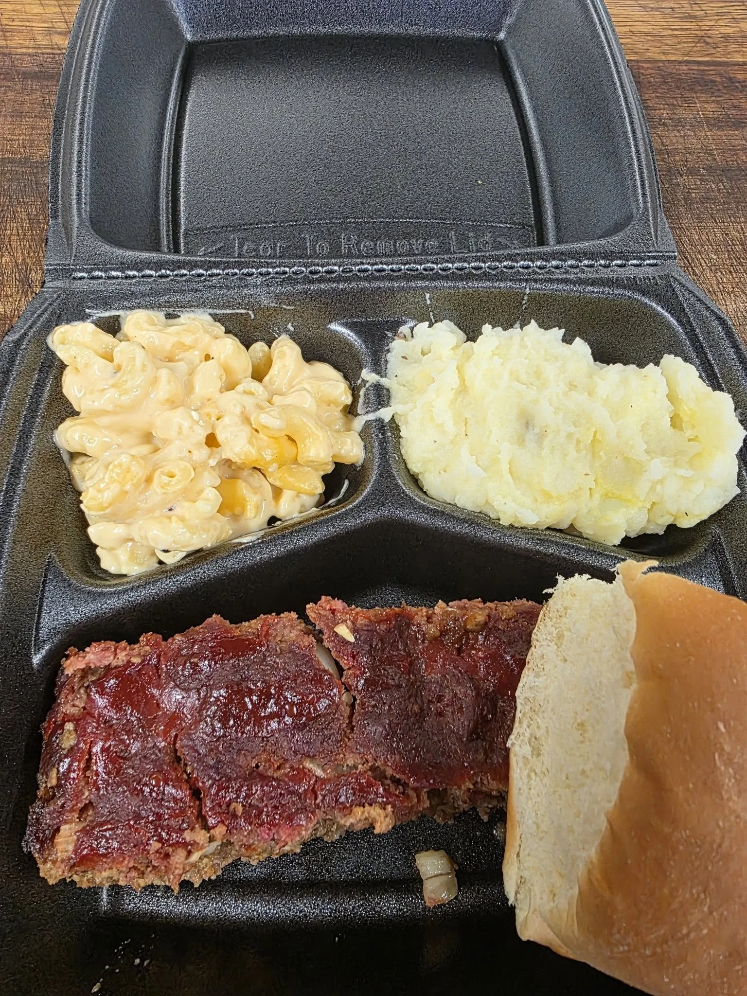 A black takeout tray with baked macaroni and cheese, mashed potatoes, meatloaf with ketchup, a bread roll, and a piece of bread with spreads on top.