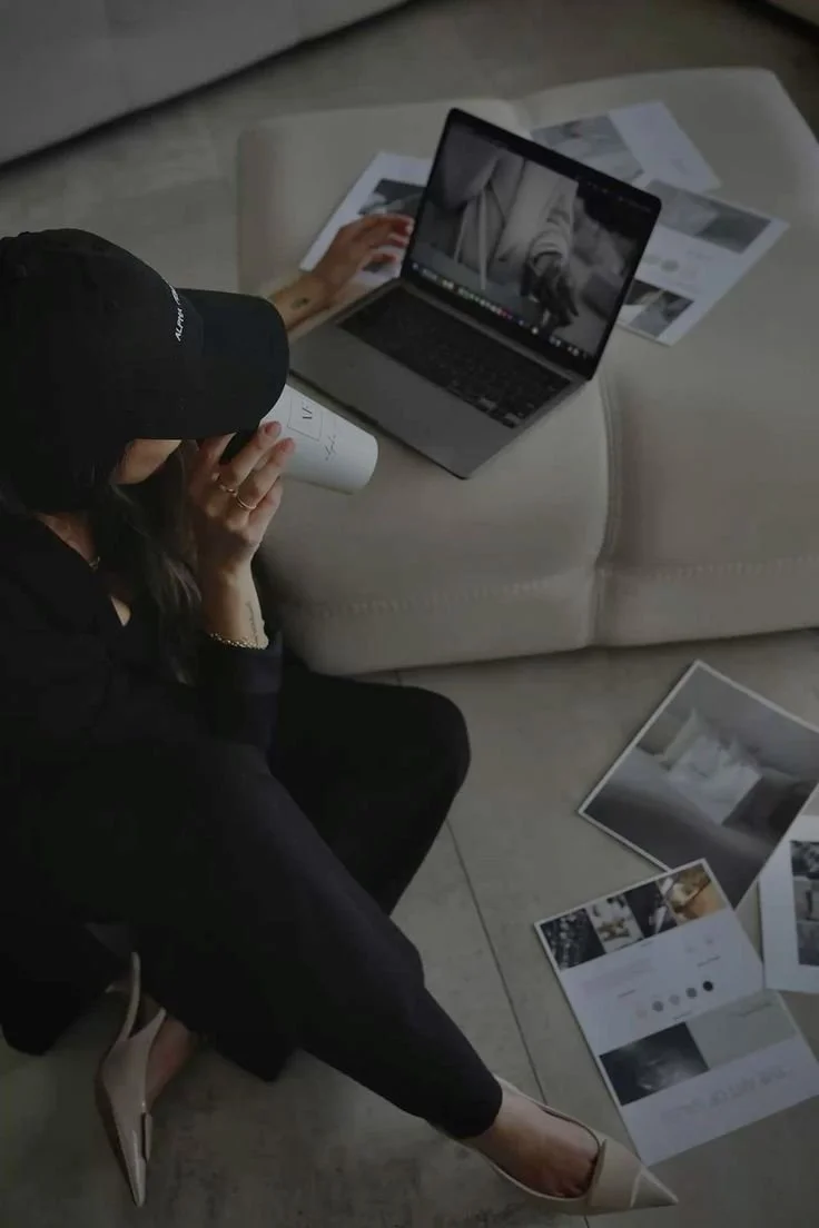 A woman sitting on the floor taking a drink while working on a laptop placed on a sofa, surrounded by printed papers and photographs.