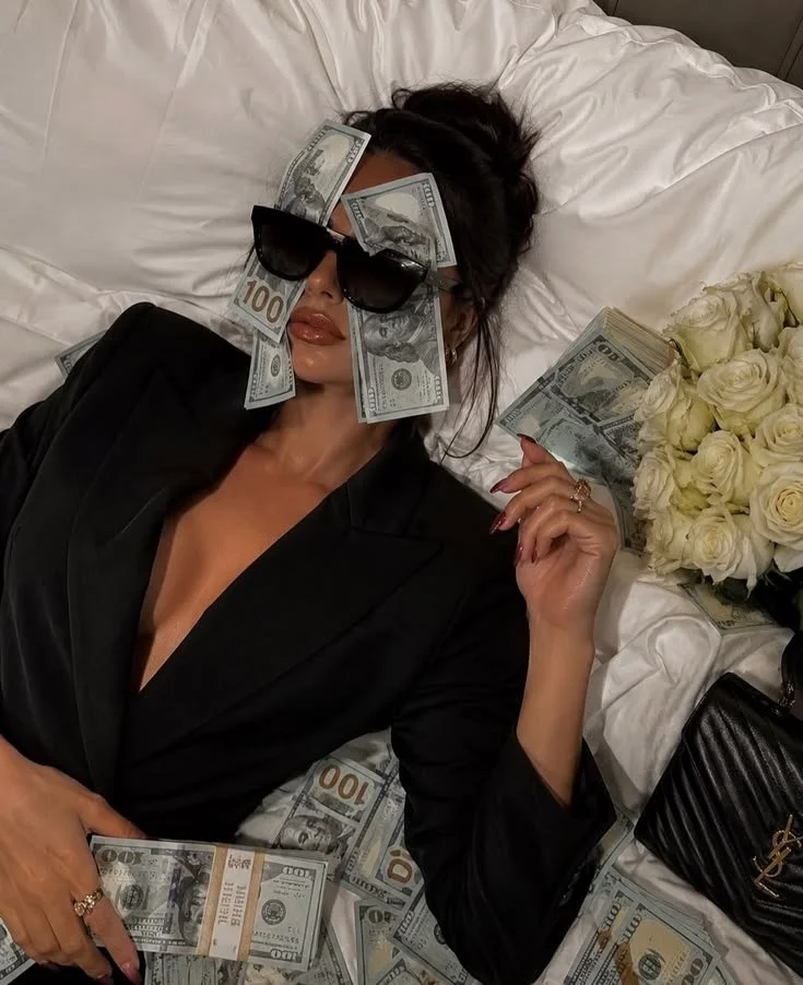 A woman lying on a bed with white sheets, wearing sunglasses and a black outfit, surrounded by hundred-dollar bills and white roses.