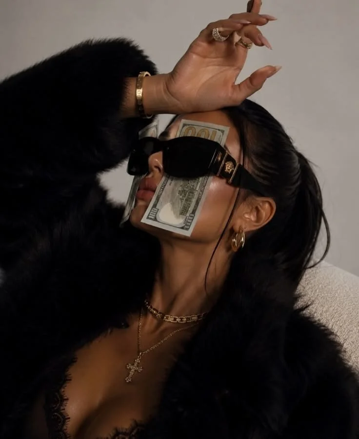 A woman with dark hair wearing black sunglasses, gold jewelry, and a black fur coat, has a hundred-dollar bill covering her eyes and part of her face, with a black cat touching her face.
