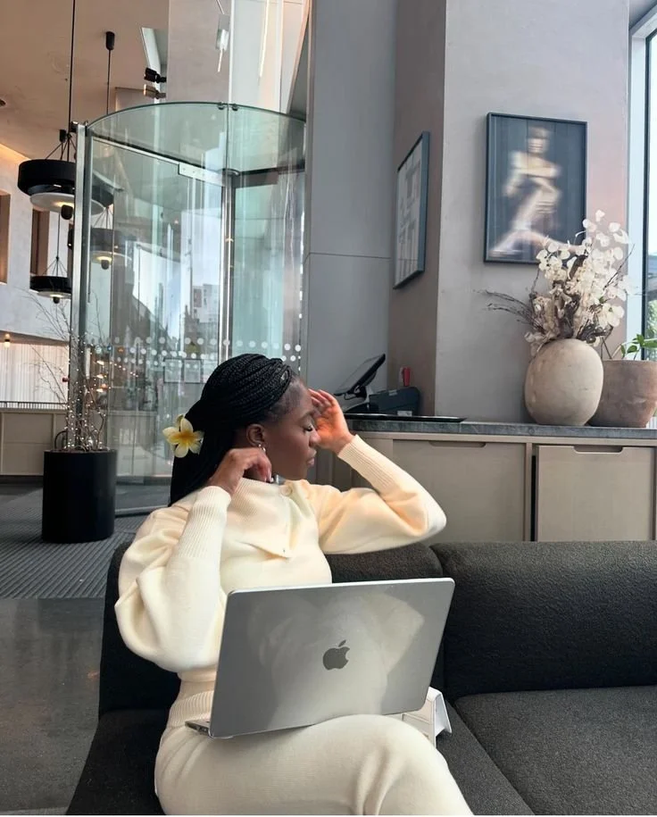 A woman sitting on a black sofa with a silver MacBook on her lap, adjusting her hair, inside a modern building with glass walls and plants.