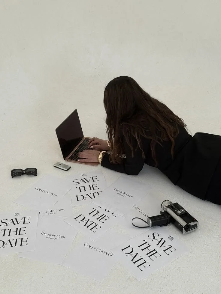 A woman is lying on her stomach on the floor working on a laptop. There are save the date business annoucement cards, a pair of sunglasses, and a camera scattered on the floor nearby.