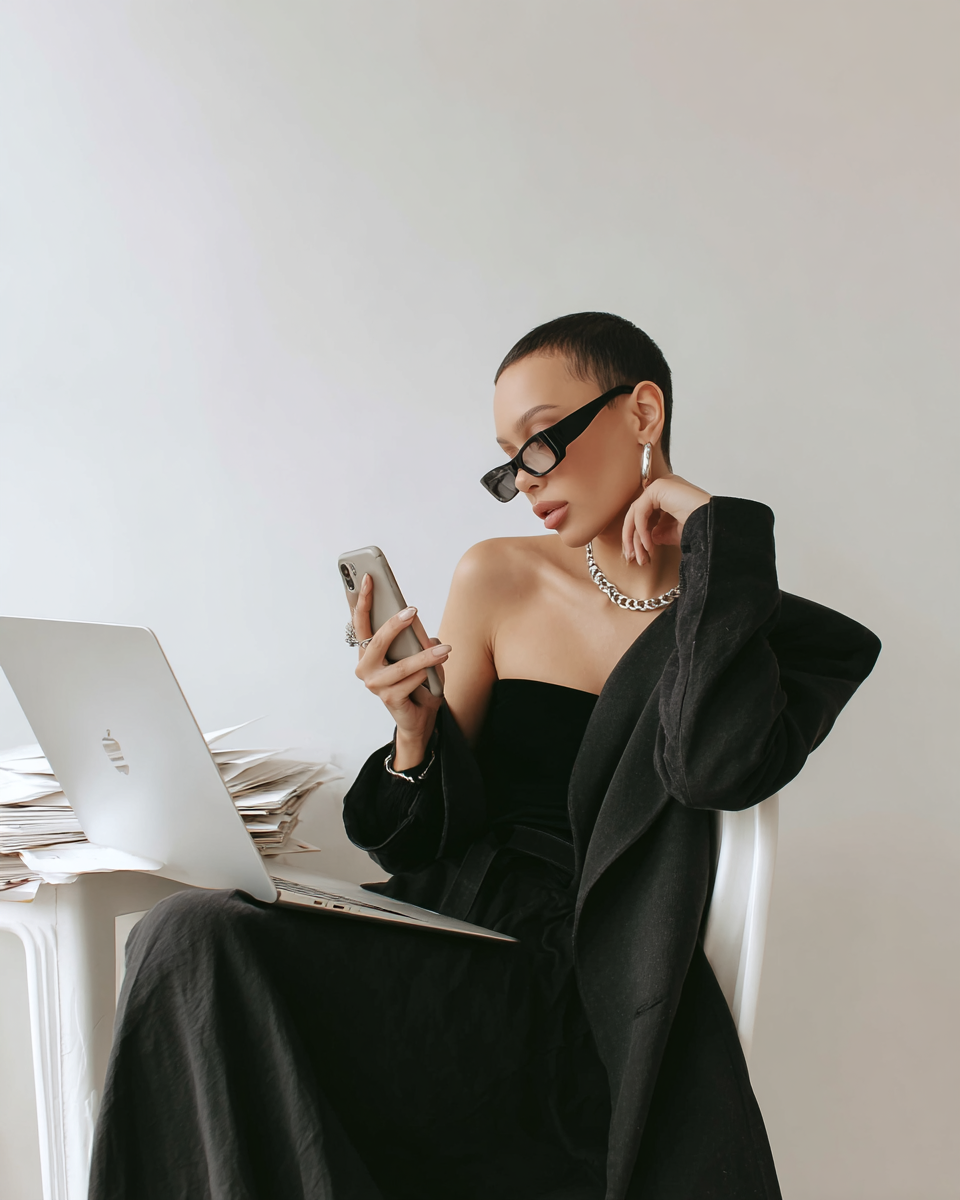 A woman with short dark hair, wearing black sunglasses, earrings, and a chain necklace, is sitting at a desk with a laptop and a large stack of papers, looking at her phone against a plain white background.