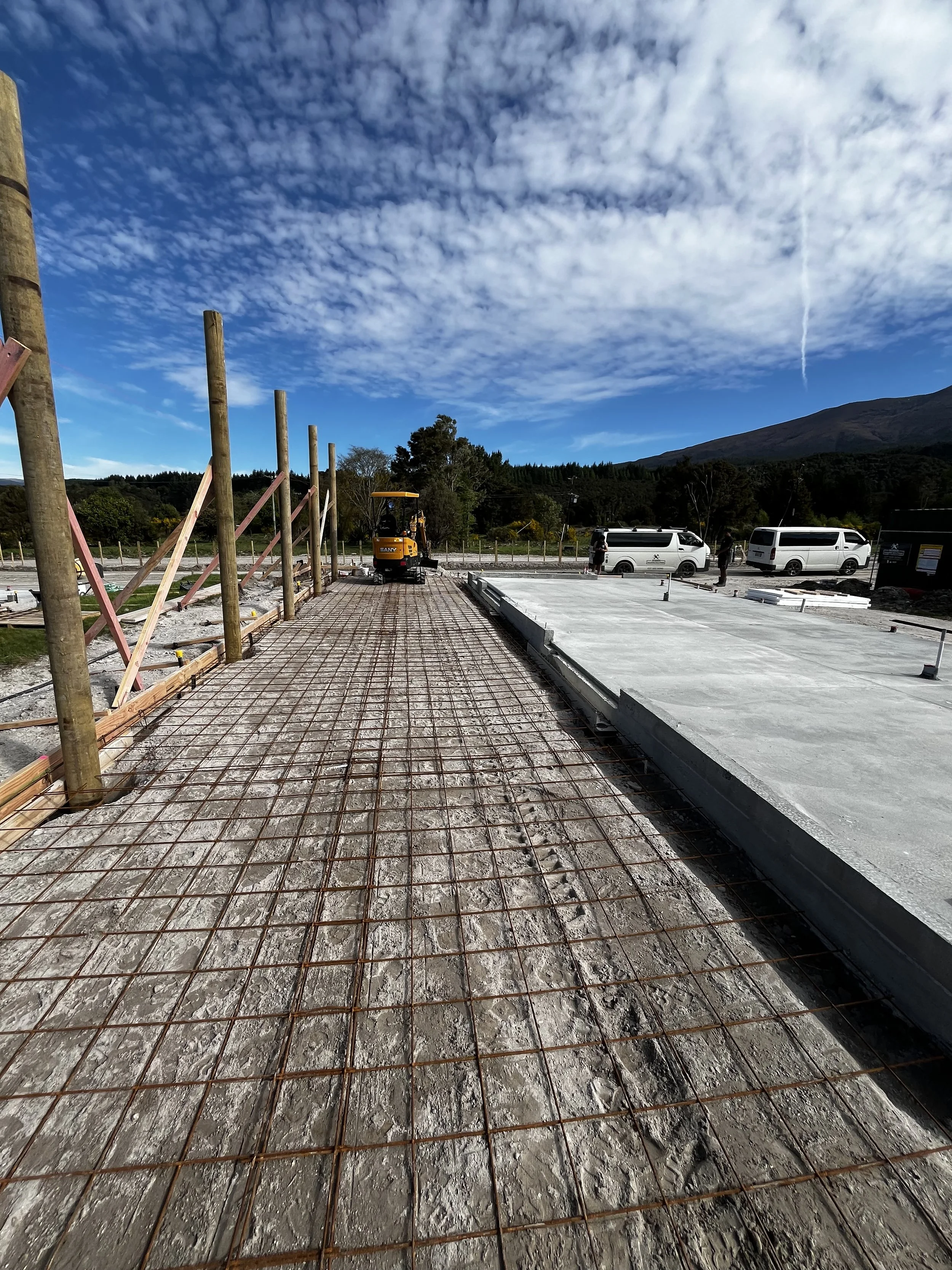 Construction site with concrete slab and metal rebar grid, wooden posts, construction equipment, and parked vans against a backdrop of trees, hills, and a partly cloudy sky.