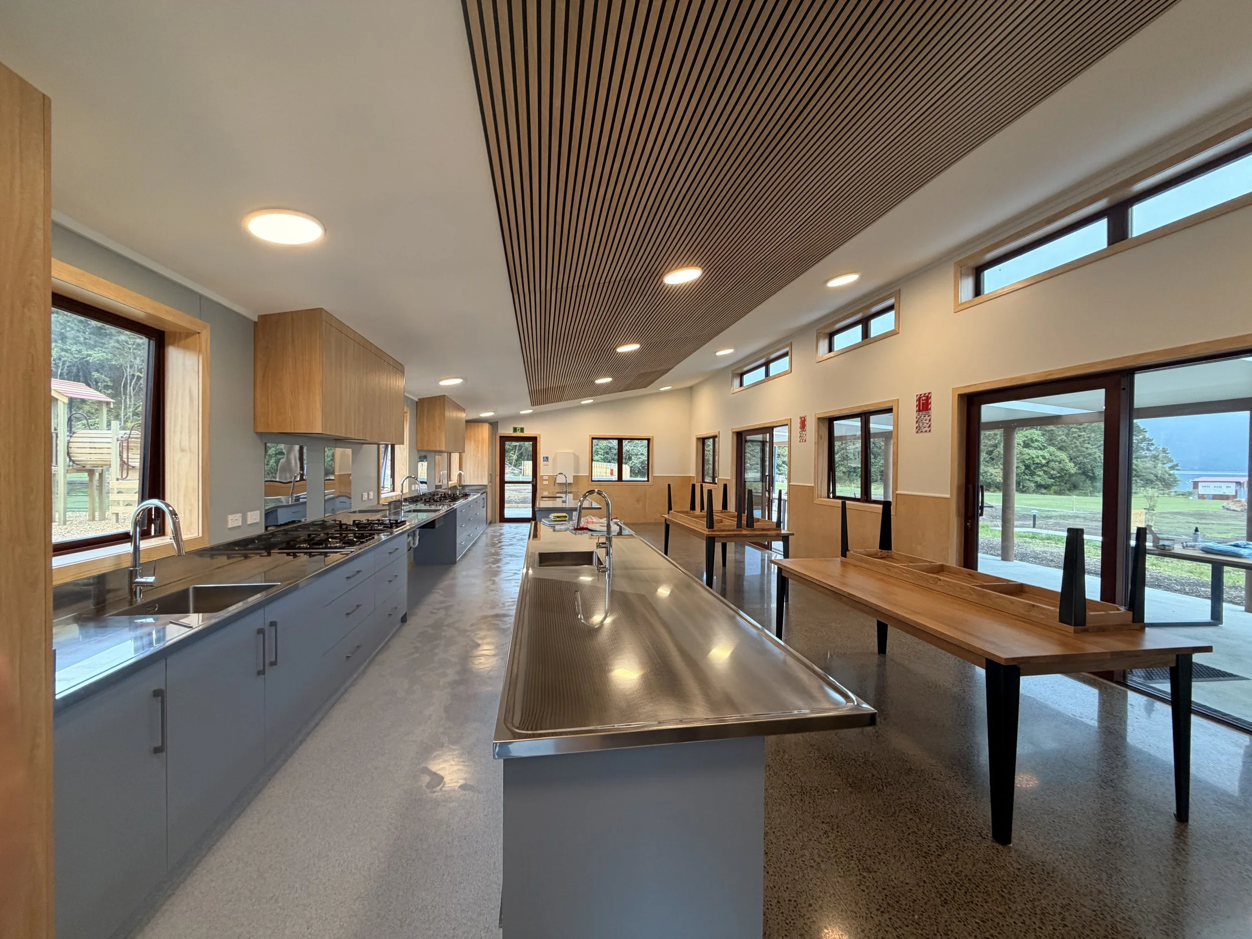 Interior view of a modern kitchen with a long countertop, multiple sinks, wooden cabinets, and ample windows showing an outdoor landscape.