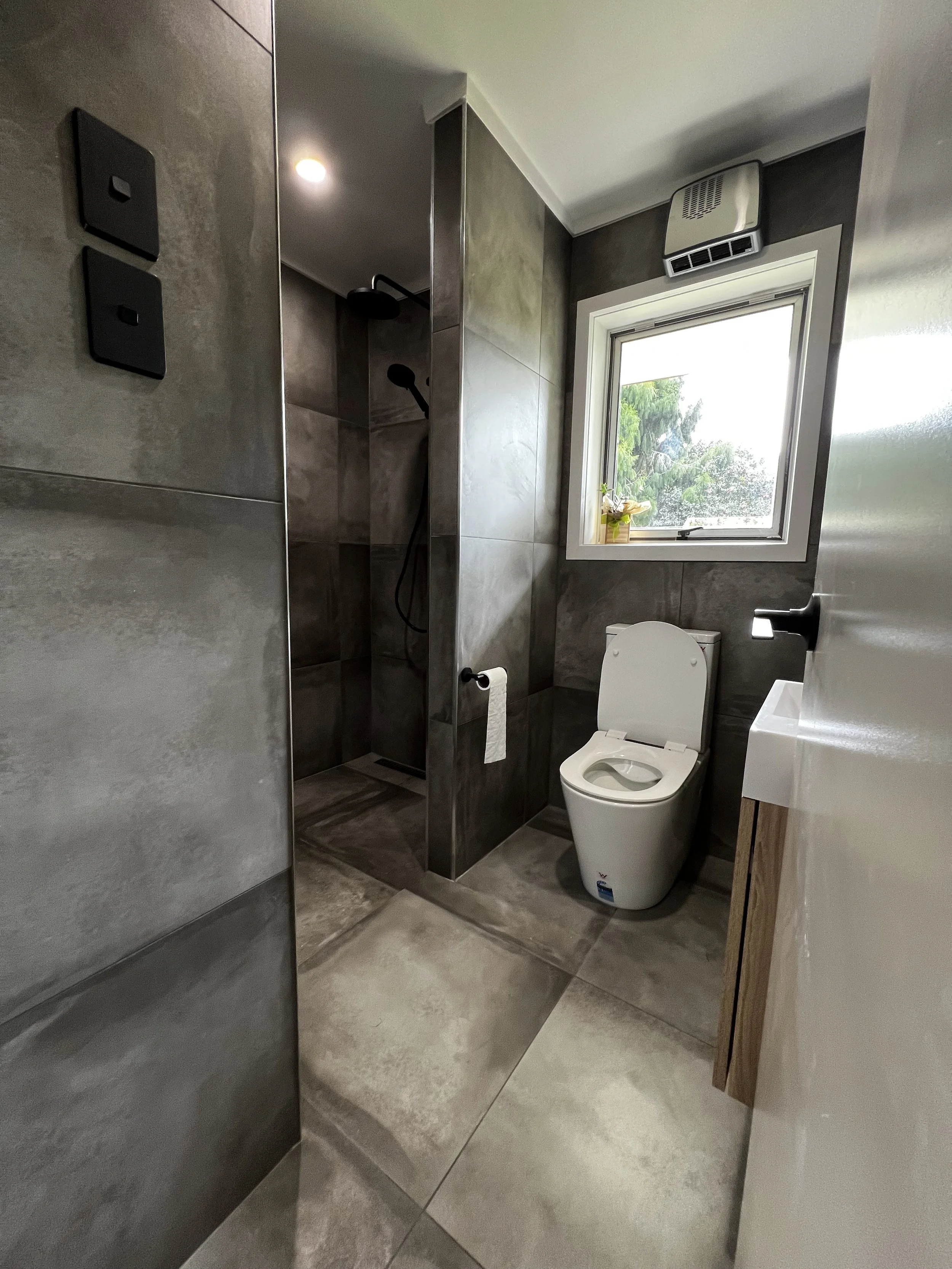 A modern bathroom with gray tile walls and floor, a toilet with an open lid, a small sink, a window with a flower in a pot, a shower area with a dark showerhead, and a ventilation fan above the window.