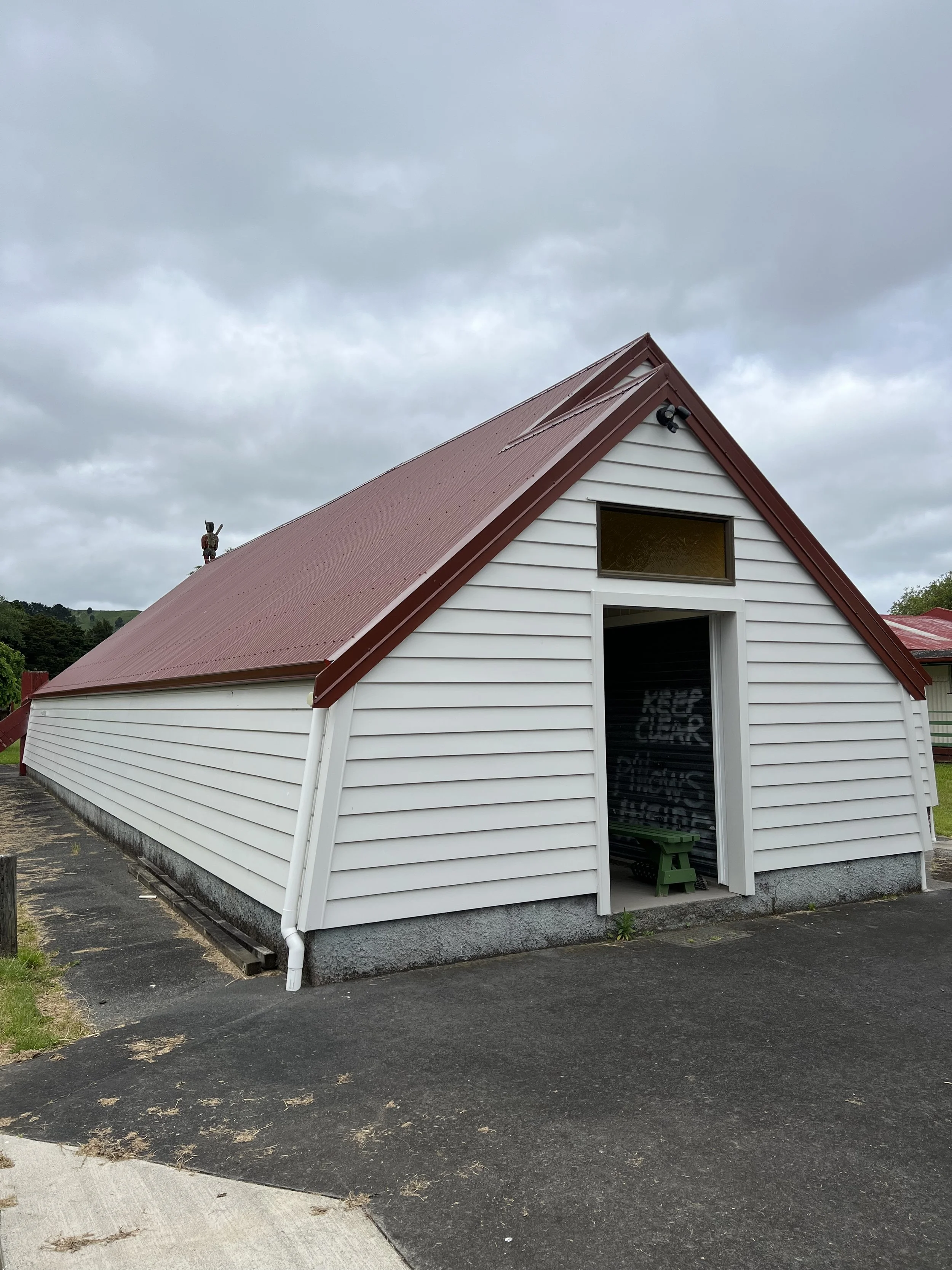 A small white building with a red metal roof, partially open door, and a window above it. A green bench is visible inside. The sky is overcast with gray clouds, and the ground is paved with asphalt and concrete sidewalk.