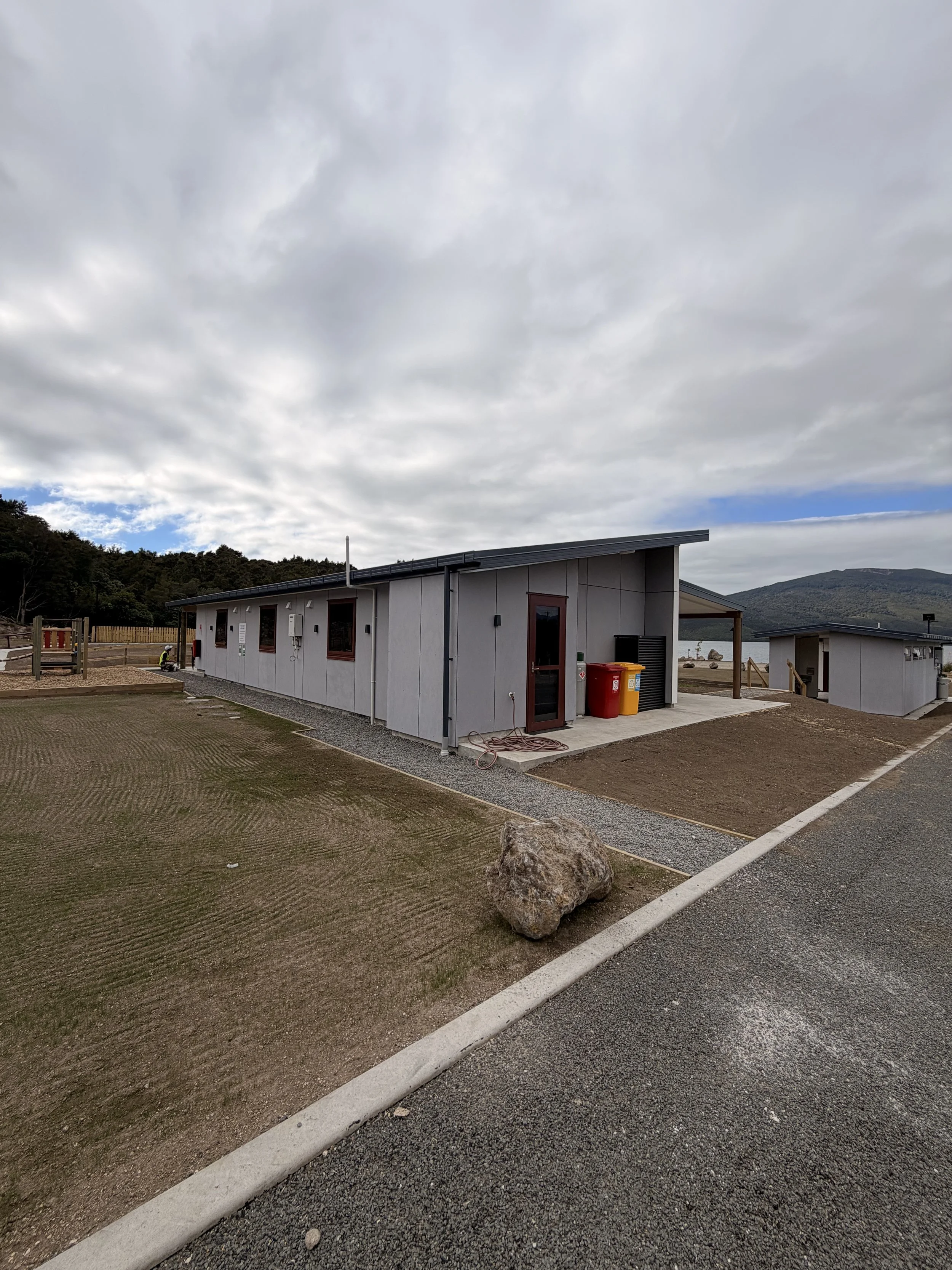 A modern building with a sloped roof, located near a lake with mountains in the background. The area around the building has gravel, grass, and some landscaping, with a large rock in the foreground.