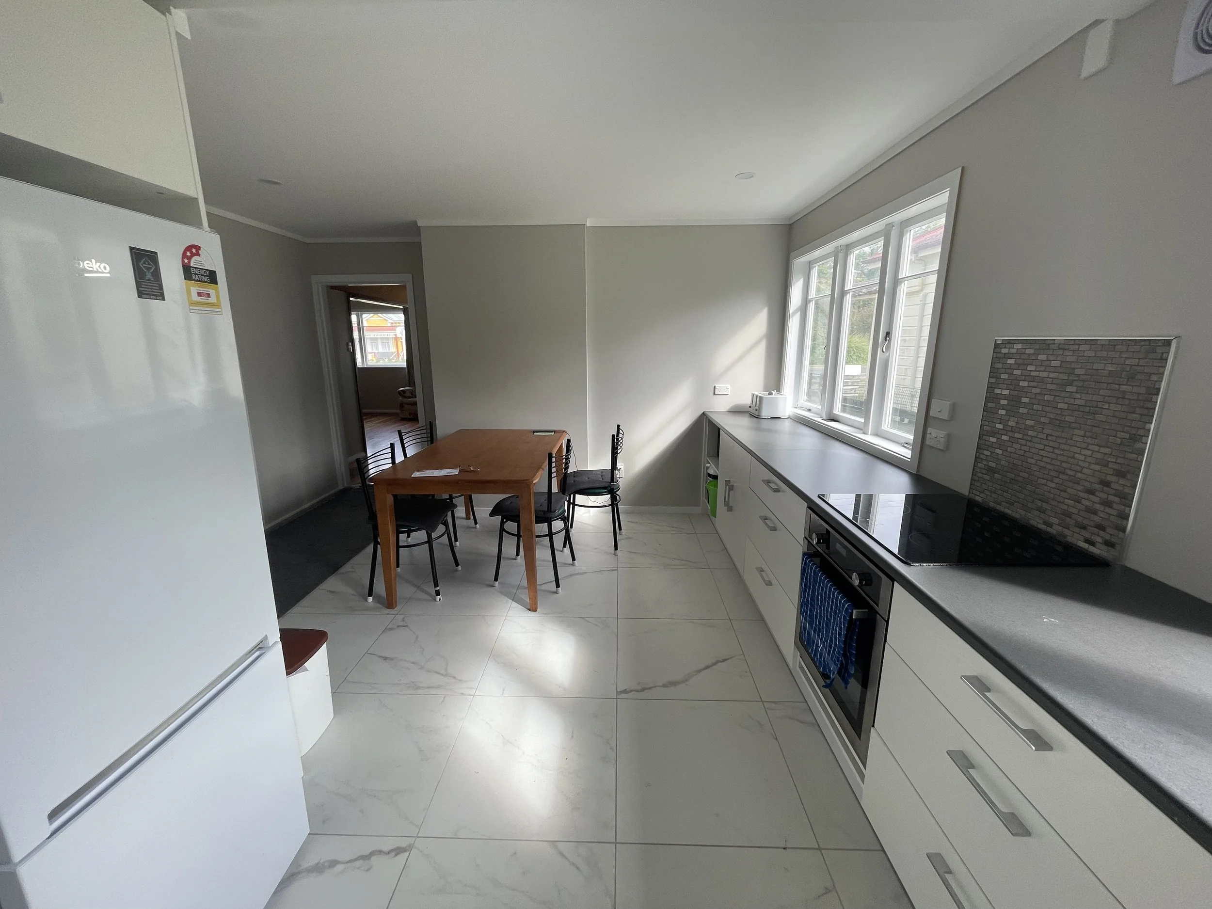 Interior of a modern kitchen and dining area with white cabinets, a black stove, a window with a view outside, and a wooden dining table with chairs.