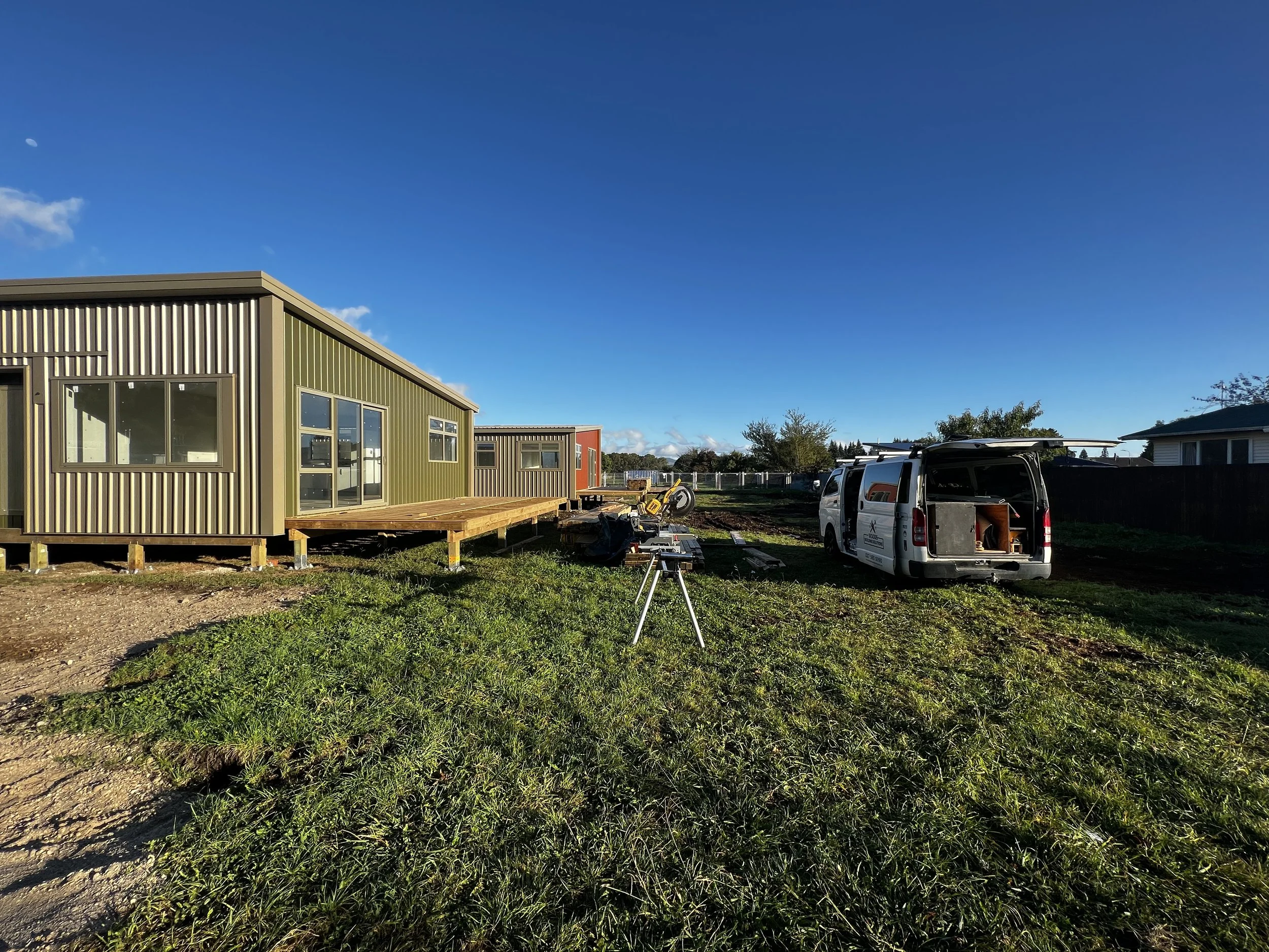Residential construction site with partially built green and red houses, construction tools, and a white van parked nearby under a blue sky.