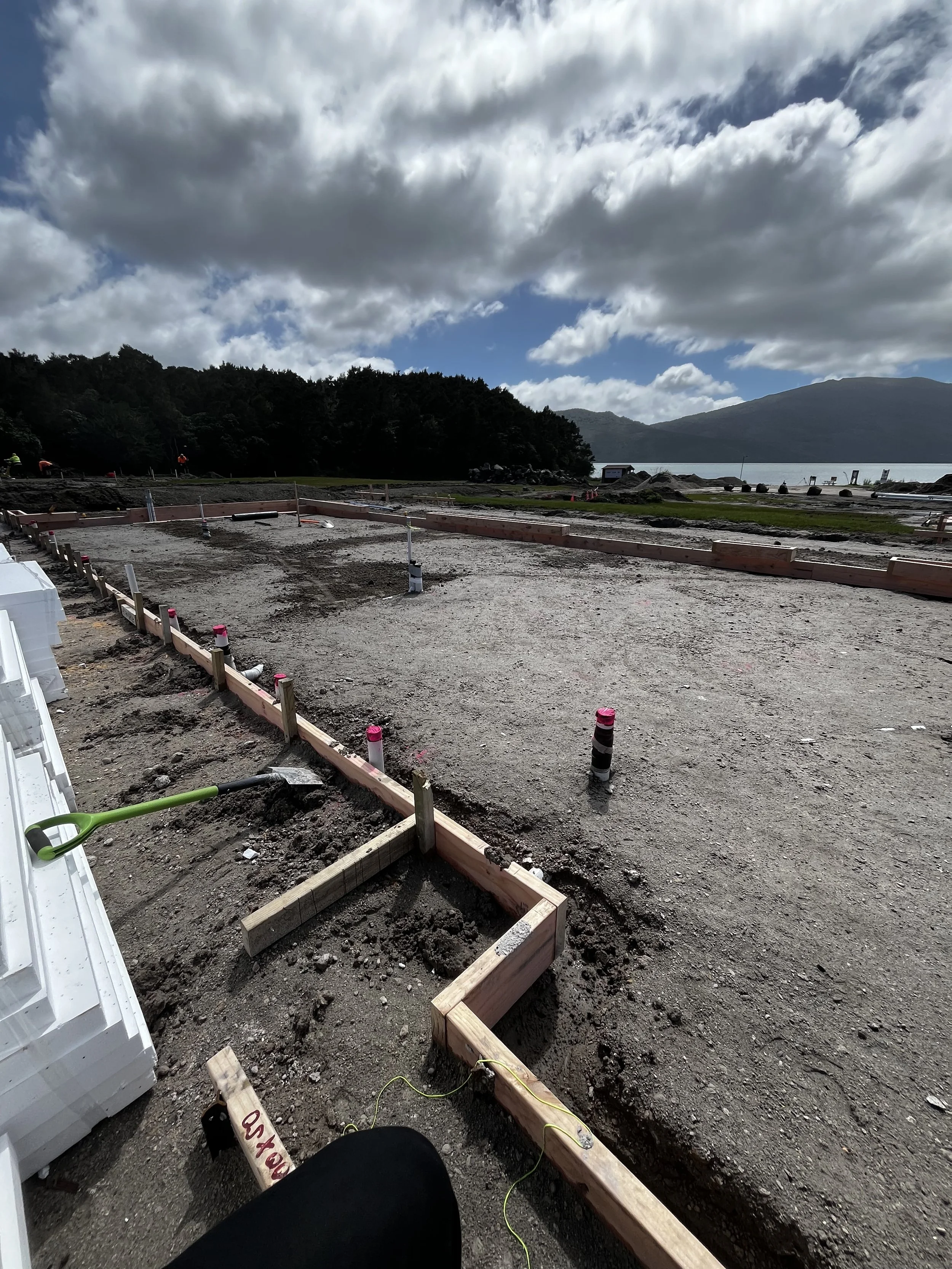 Construction site with framing in progress, gravel ground, and a scenic view of water, mountains, and cloudy sky in the background.