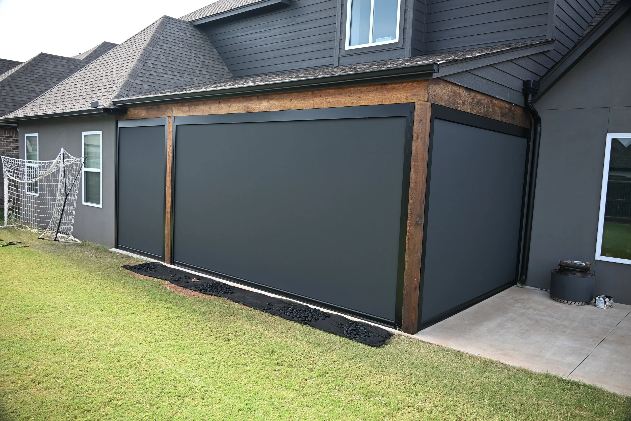 The image shows the back of a house with a newly installed or being installed gray sliding door or window shutter. There is a grassy lawn in the foreground and a patio area with concrete flooring. A soccer goal net is leaning against the wall near tw