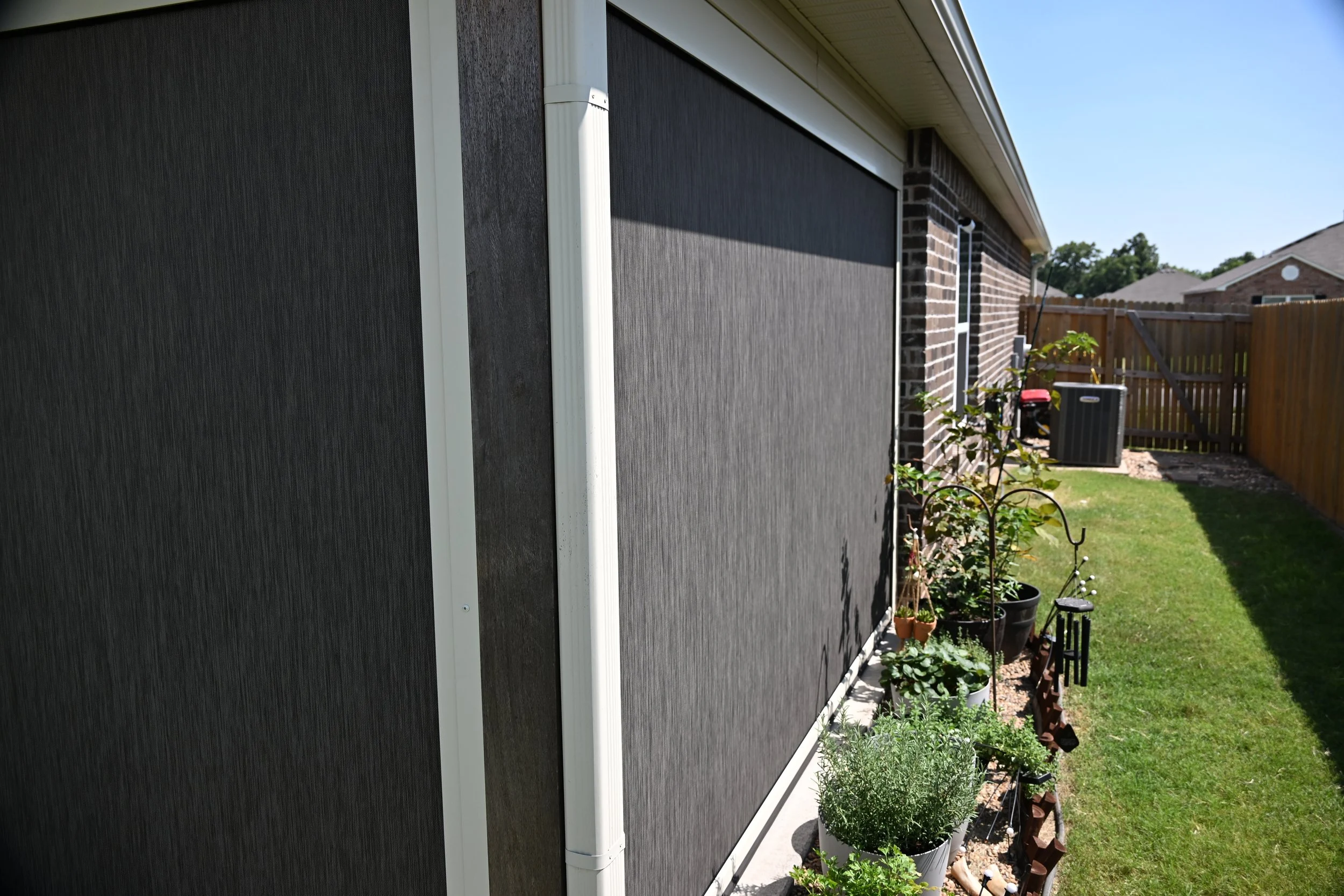 Side view of a house with a black exterior wall, white trim, and a brick wall, backyard with plants, fence, and lawn under a clear blue sky.