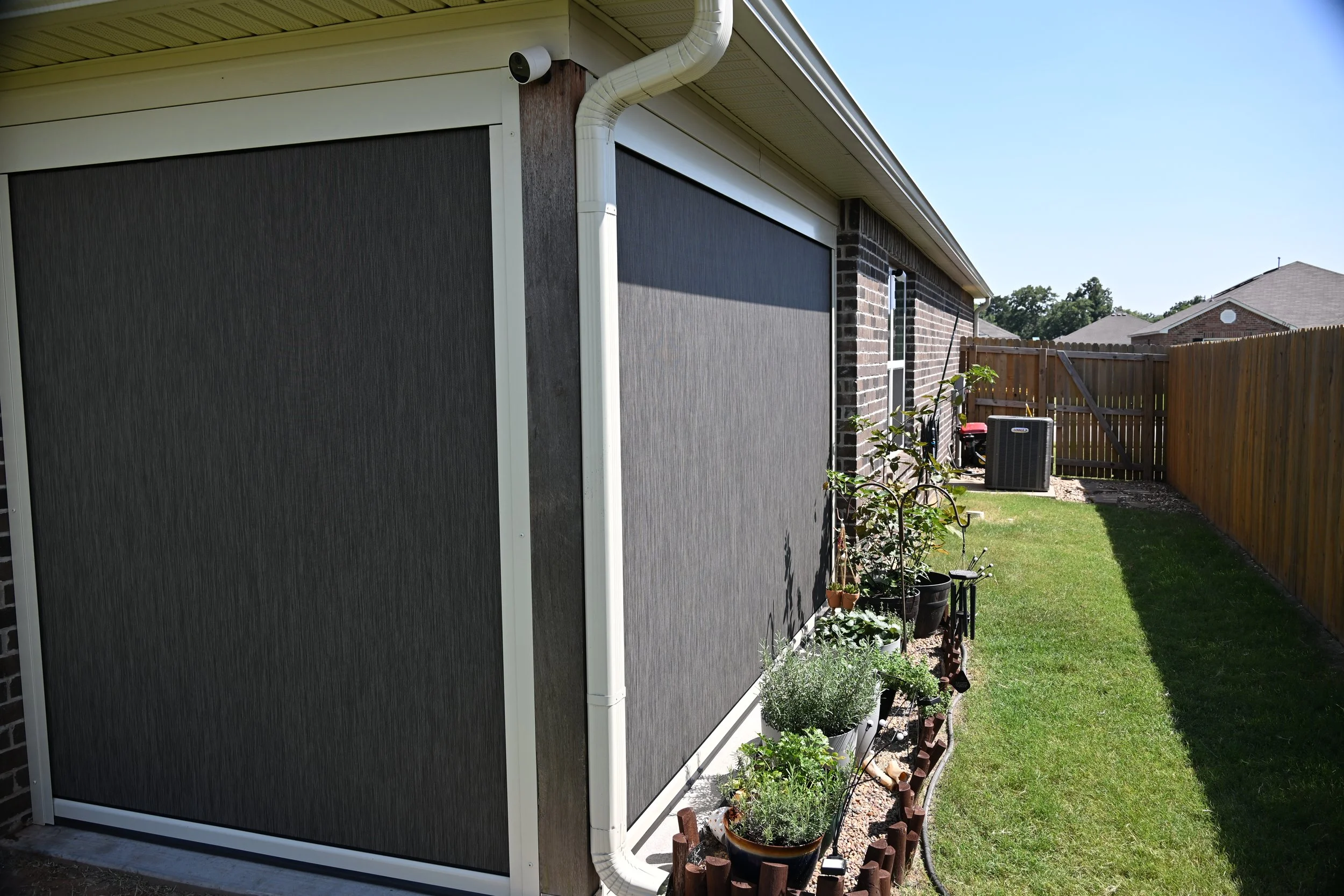 A backyard with a brick house, a wooden fence, a small garden with potted plants, and an air conditioning unit next to the fence, under a clear blue sky.