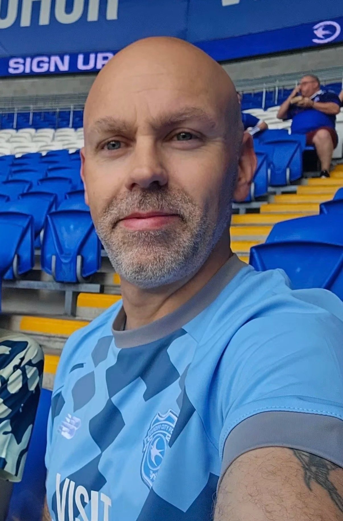 A man with a bald head, gray beard, and blue eyes taking a selfie in a stadium. He is wearing a blue sports jersey with a logo and the word 'VISIT' on it. In the background, blue and yellow stadium seats and a man sitting on the stairs talking on a phone are visible.