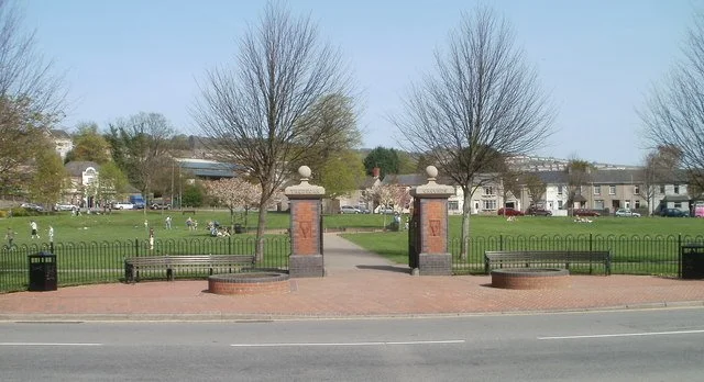 A park entrance with brick pillars and iron gates, with trees and houses in the background.