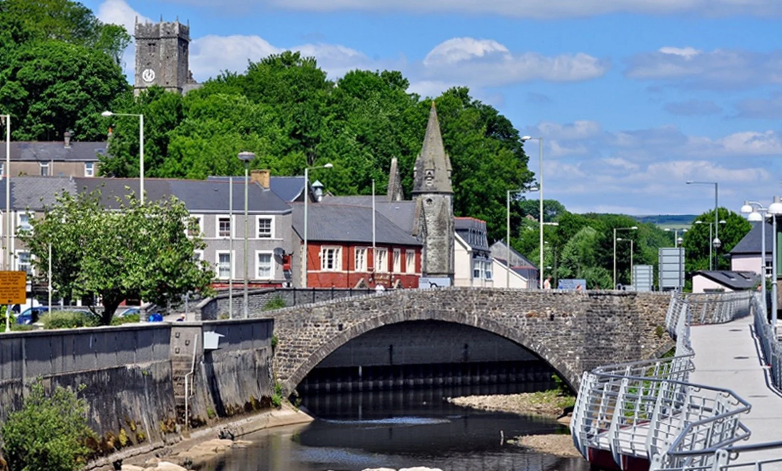 A scenic view of a stone bridge crossing a small river with historic buildings and church steeples behind, green trees, blue sky, and clouds.
