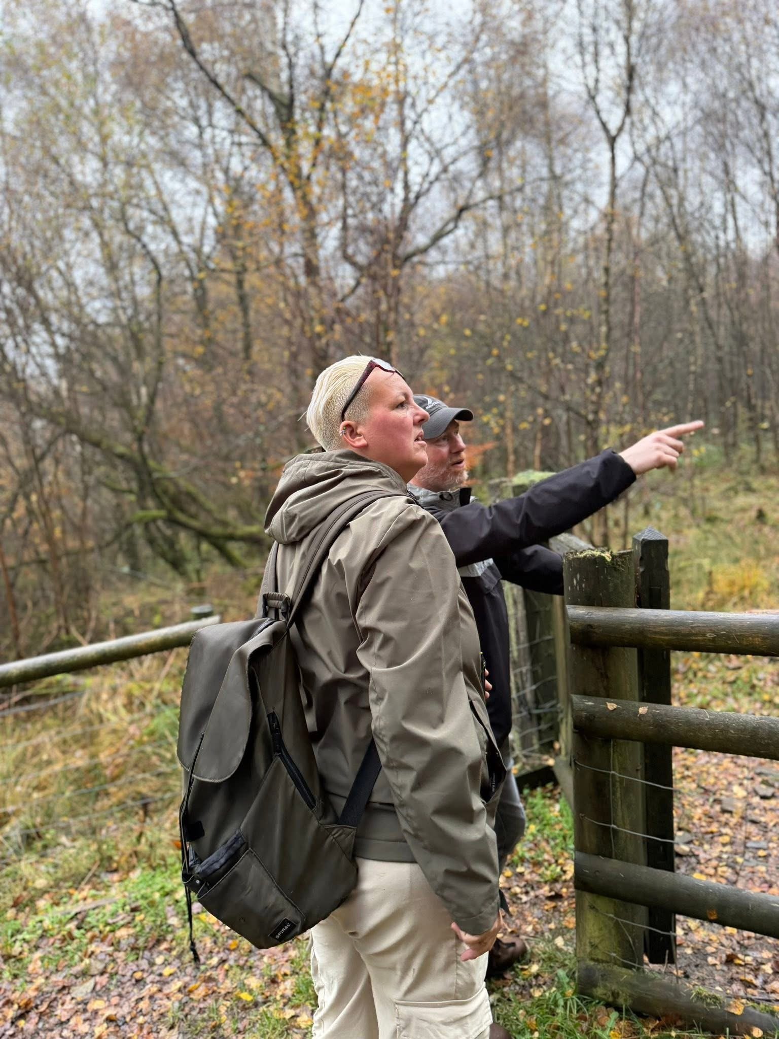 Two people standing outdoors in a forested area with fall foliage, one of them pointing at something.