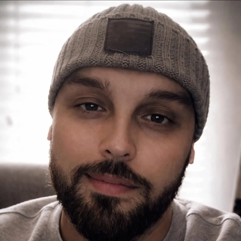 Close-up of a man with a beard and mustache, wearing a beige knit beanie with a black patch on the front, indoors with window blinds in the background.