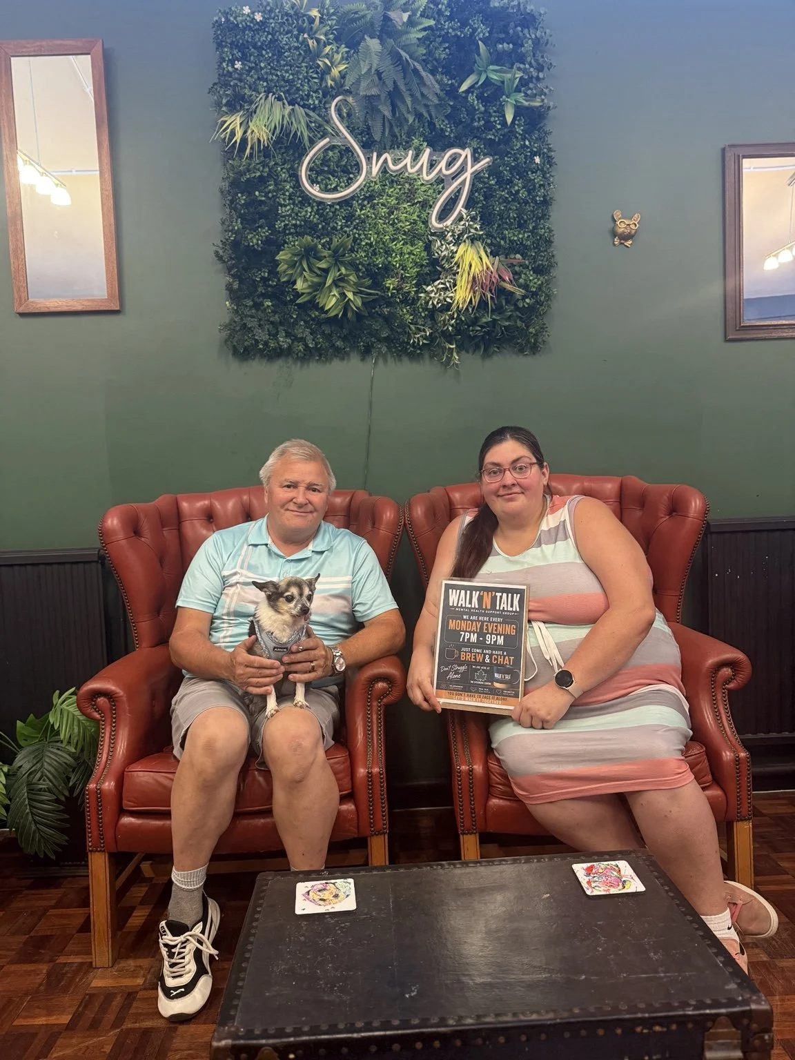 A man and woman sit on red leather armchairs in a cozy indoor setting with green walls and greenery decor. The man holds a small dog, and the woman holds a sign promoting a weekly chat event. There are two mirrors on the walls and a small table in front with decorative coasters.